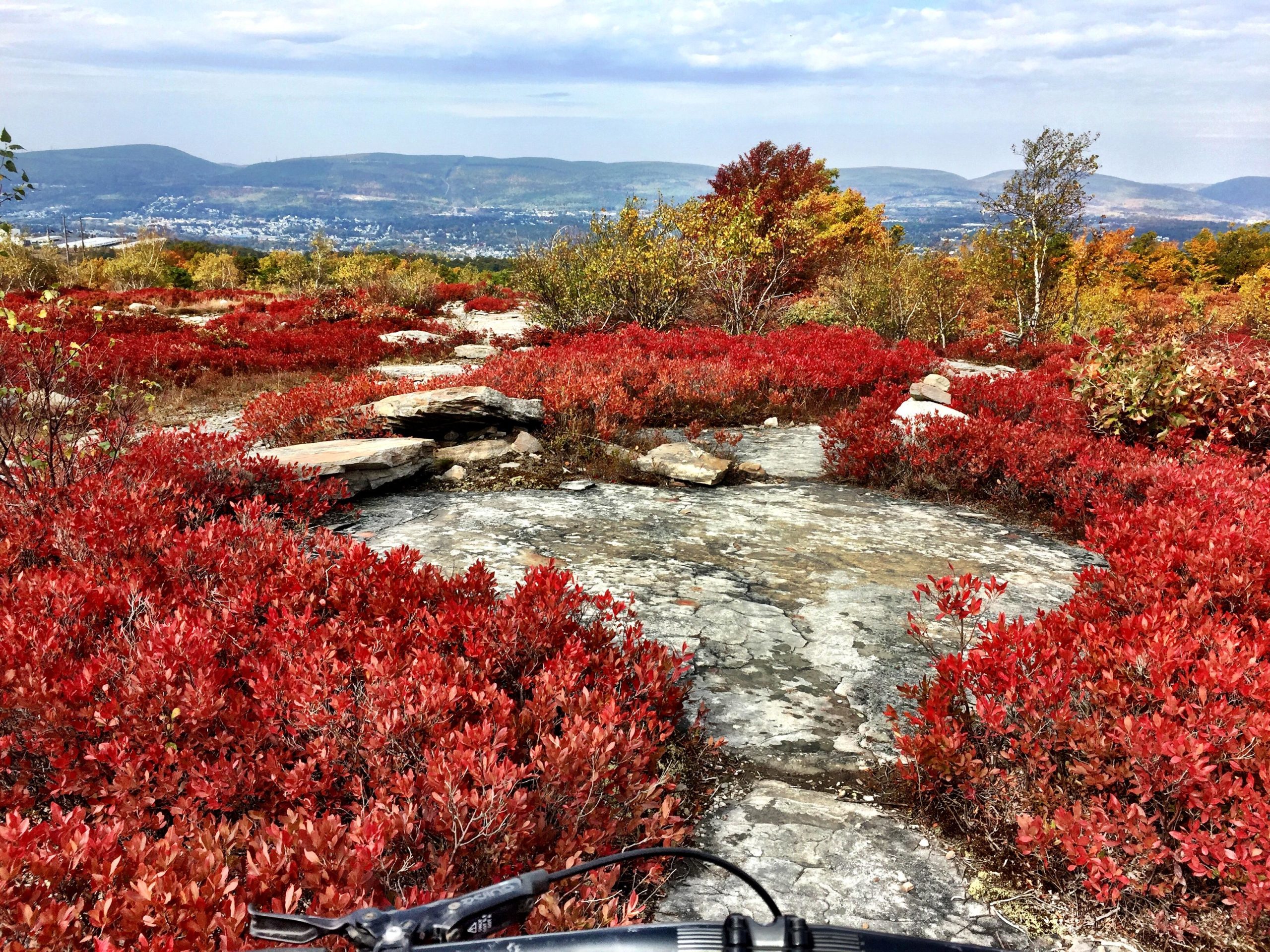 A scenic view from a rocky hillside, featuring vibrant red foliage in the foreground and rolling hills in the background under a partly cloudy sky. The image captures the beauty of autumn with layered vegetation and rocky surfaces leading into the distance. Blueberry mountain bike trail.