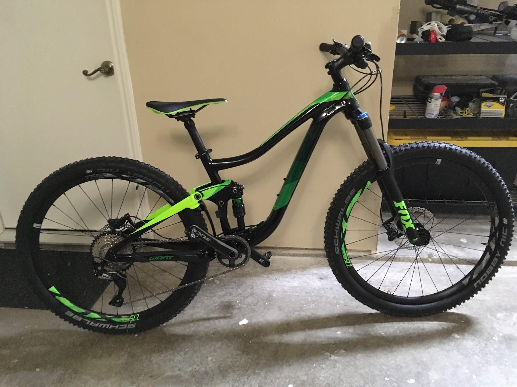Giant Trance 2: A black and green mountain bike leaning against a wall, featuring thick tires and a modern frame design. The bike has a visible suspension system and is positioned on a concrete floor with a storage area in the background.