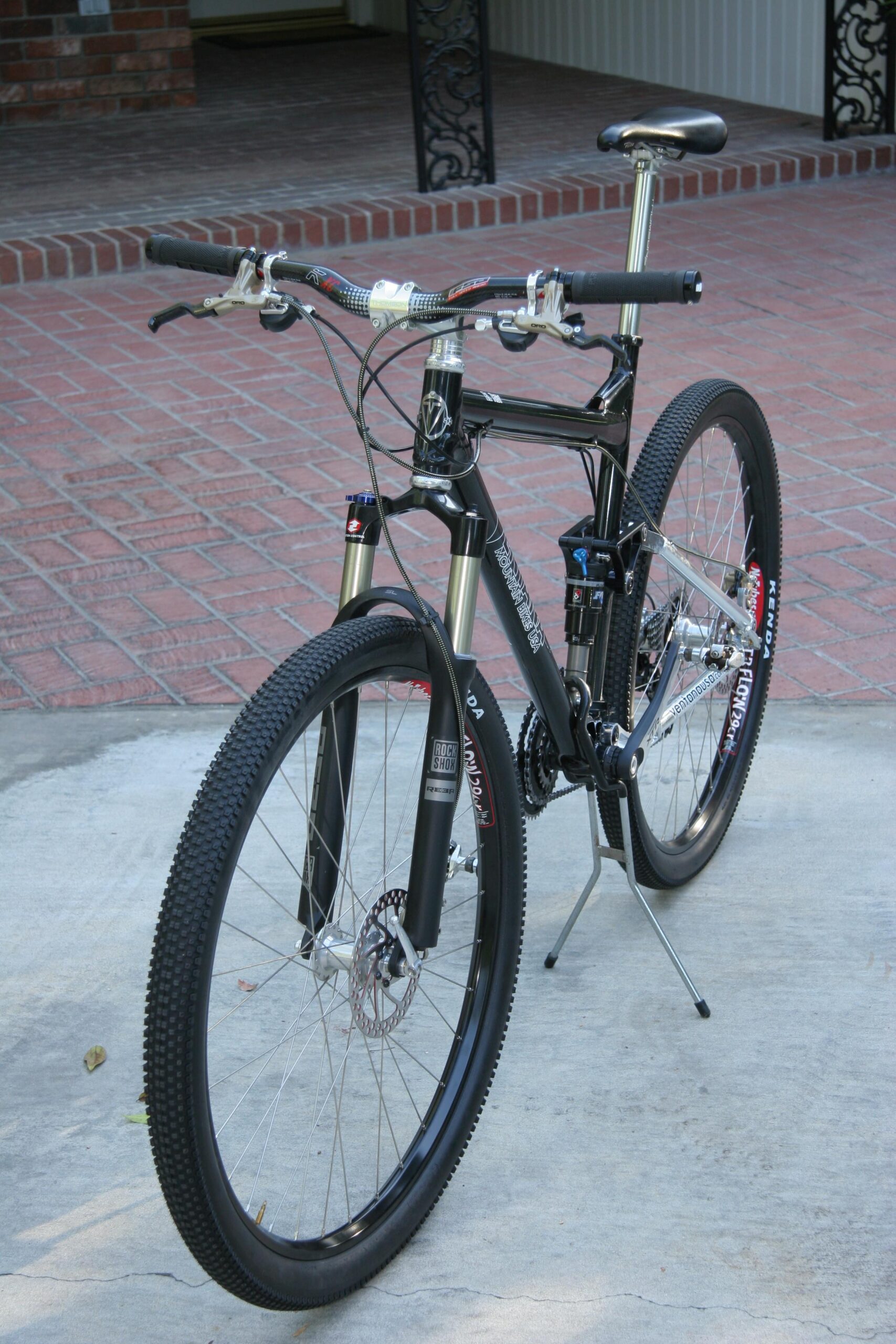Ventana El Ray El Ray: A black mountain bike with a front suspension system, standing upright on a concrete surface. The handlebars feature protective grips, and the tires have a pronounced tread pattern suitable for off-road terrain. The bike is positioned near a brick pathway and a structure with a decorative iron feature in the background.
