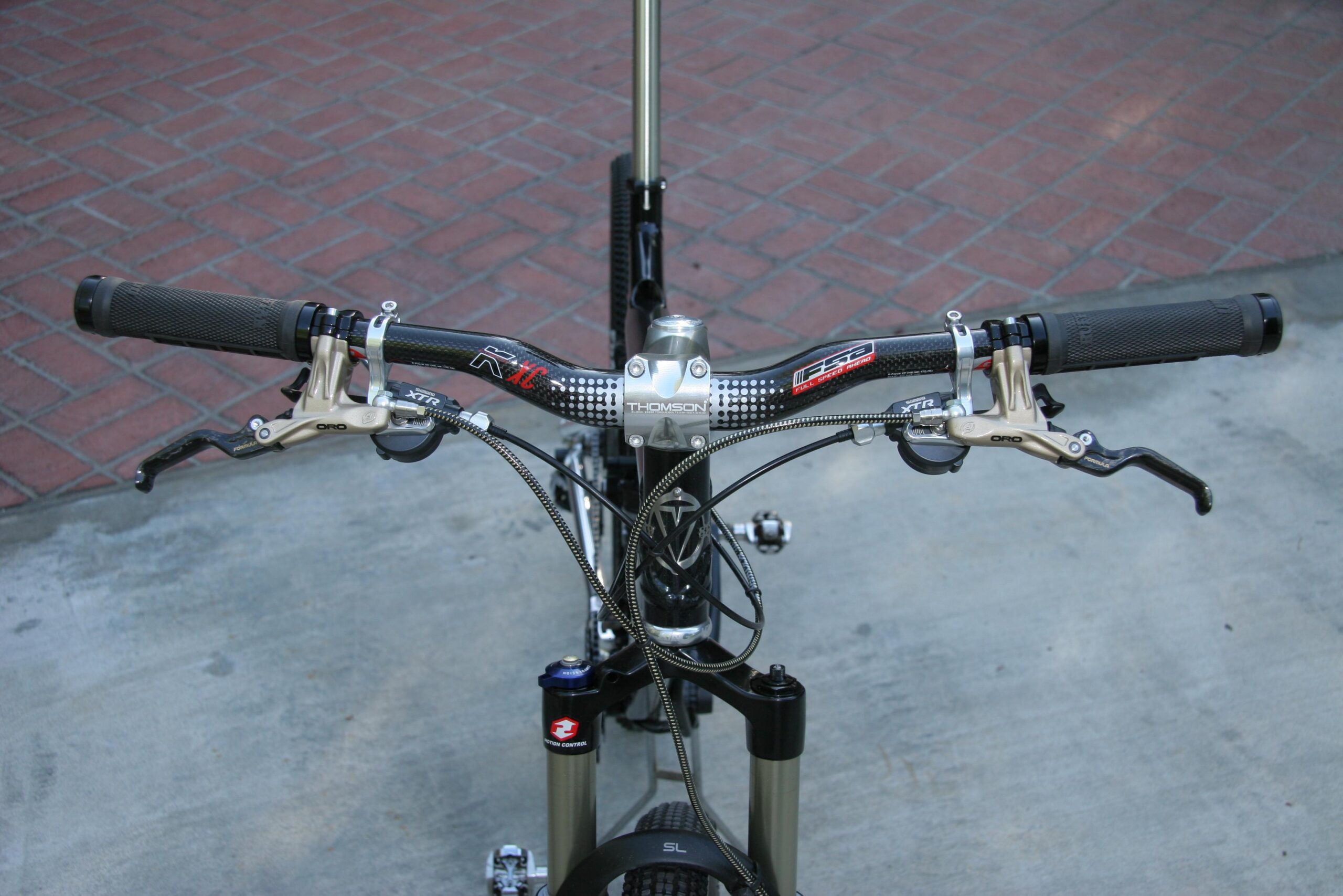 Ventana El Ray El Ray: Close-up view of a bicycle handlebar setup, featuring textured grips, brake levers, and a prominent stem. The handlebars are mounted on a fork with visible suspension components, positioned over a textured concrete surface and surrounded by a brick pattern in the background.