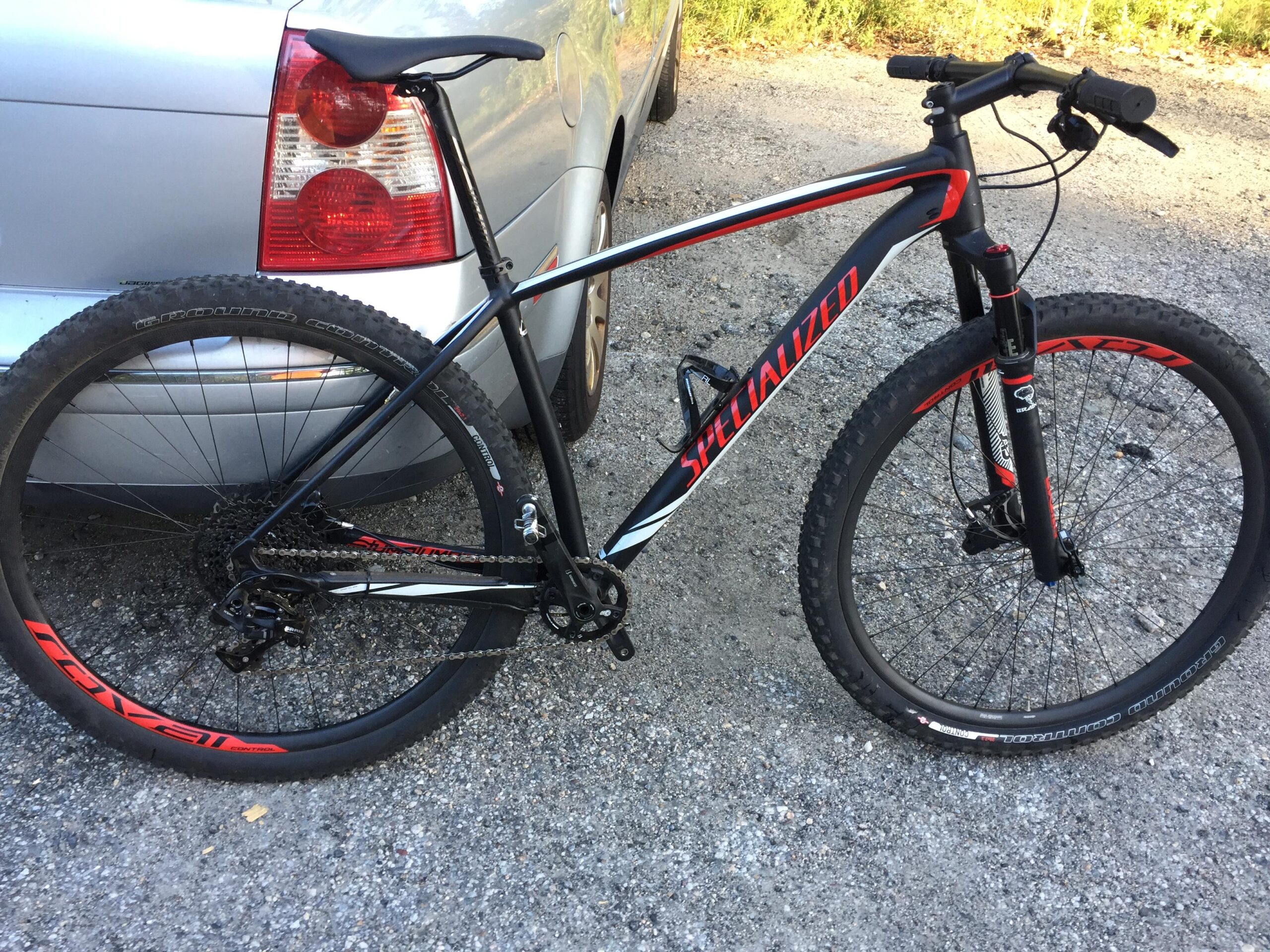 Specialized Stumpjumper: An angled view of a black and red Specialized mountain bike resting against a silver car. The bike features large tires, a prominent front suspension fork, and a gear setup visible near the rear wheel, set on a gravel surface.