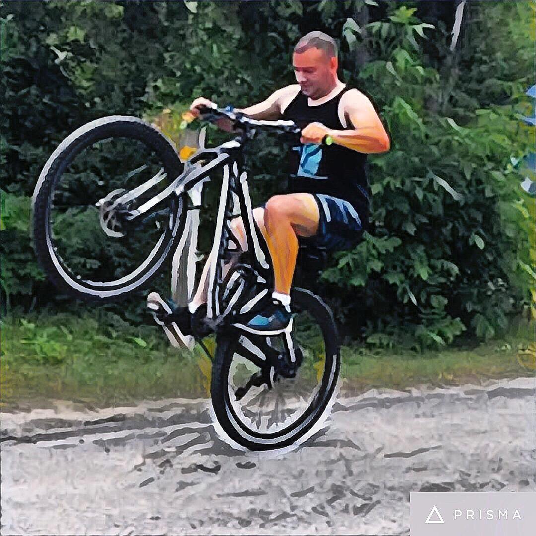 Devinci Troy: A person performing a wheelie on a mountain bike, demonstrating balance and skill, with a background of greenery and a dirt path.