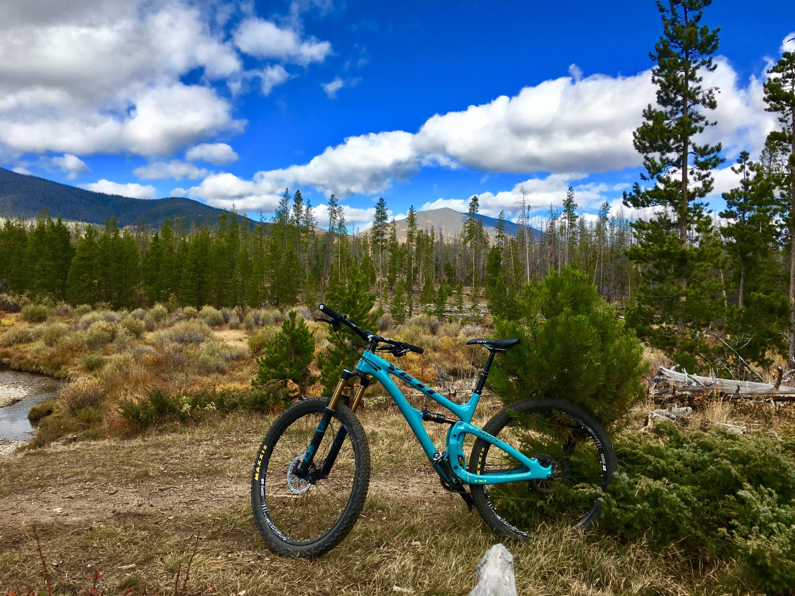 Yeti SB4.5c: A mountain bike stands on a dirt path in a scenic outdoor landscape, surrounded by greenery and shrubs. In the background, there are tall pine trees and distant mountains under a partly cloudy blue sky. A small stream runs nearby, enhancing the picturesque natural setting.