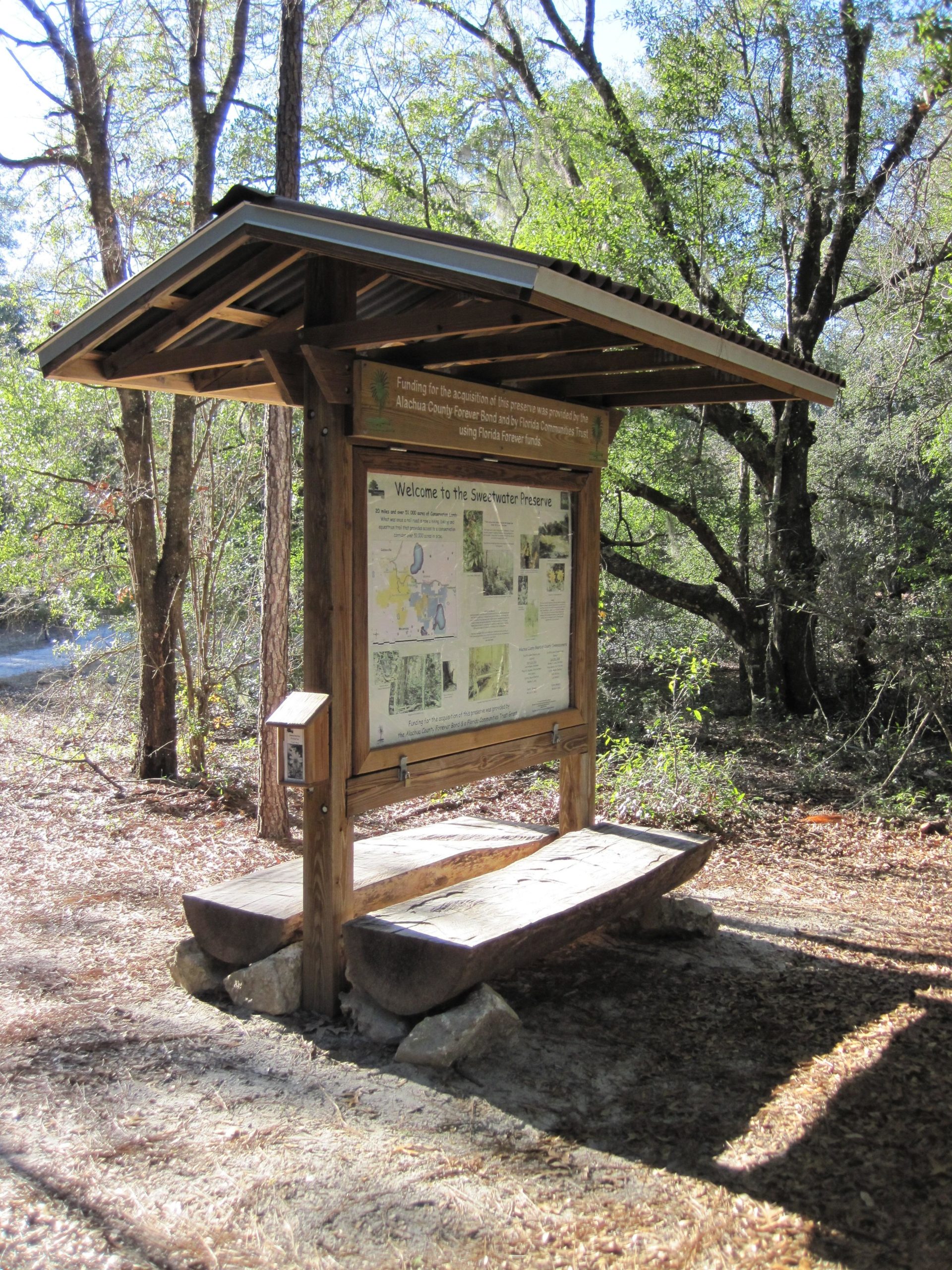 A wooden information kiosk with a sloped roof stands along a forest trail, surrounded by trees. The kiosk displays a welcome sign for the Sweetwater Preserve and includes a detailed map and information about the area. There is a wooden bench beneath the kiosk, and the ground is covered with leaves and forest debris. Sweetwater Preserve mountain bike trail.