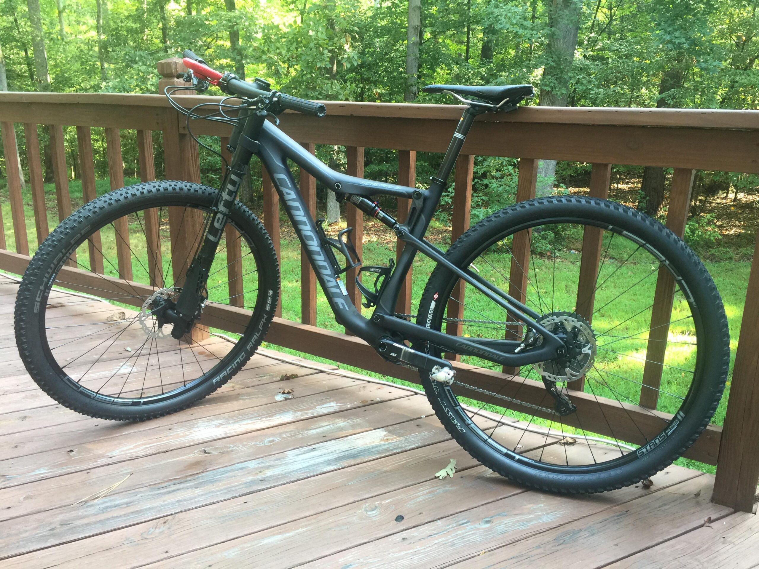 Cannondale Scalpel Carbon 3: A black mountain bike is leaning against a wooden deck railing, surrounded by green trees and grass. The bike features wide tires and a sleek frame, with visible brand markings. Natural light filters through the leaves, creating a serene outdoor setting.