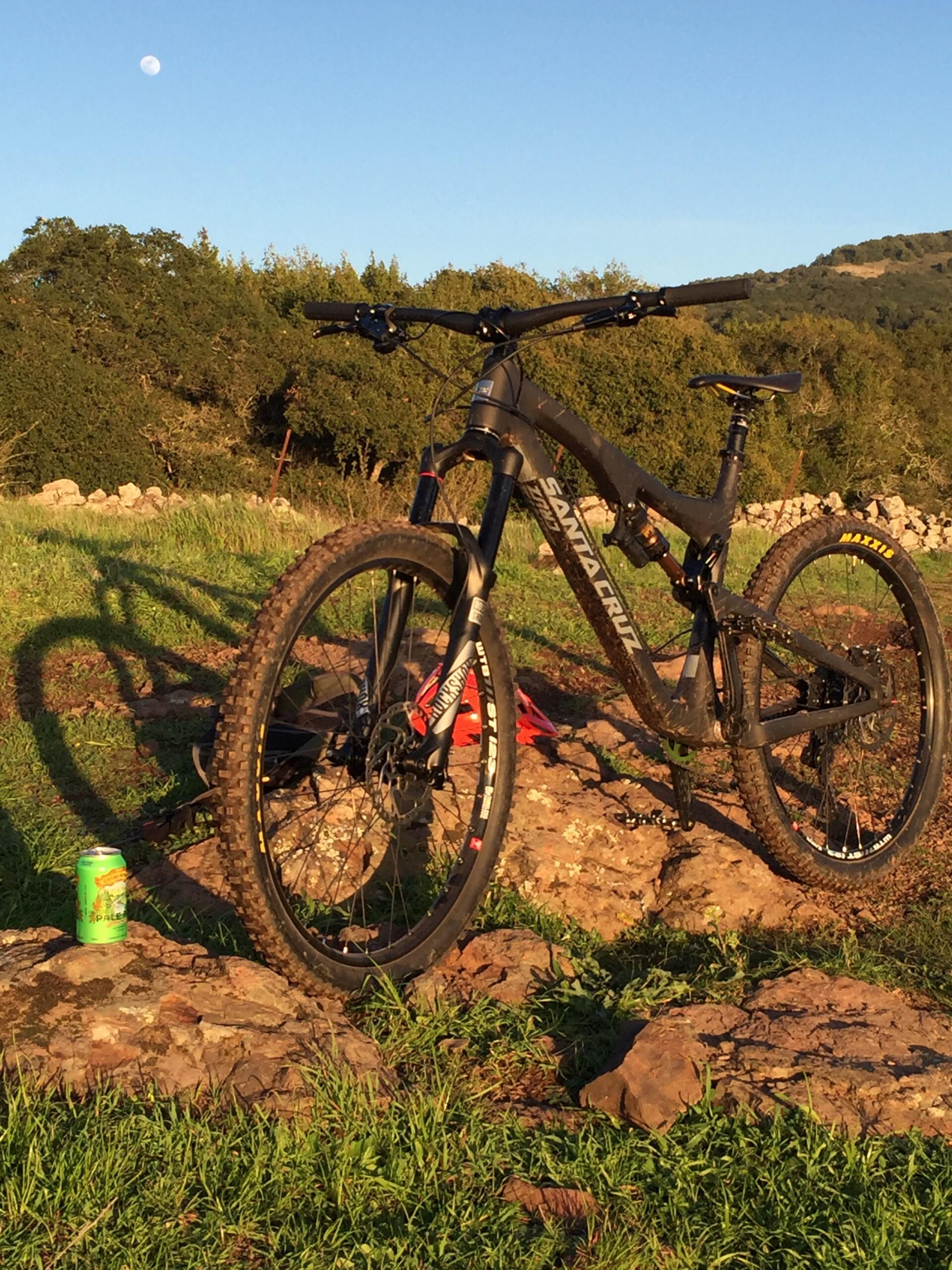 Santa Cruz Bronson 2 CC: A mountain bike resting on a rocky surface with lush green grass, under a clear blue sky. In the background, a full moon is visible, and the landscape features trees and hills. A green can of beverage sits nearby on the rocks.