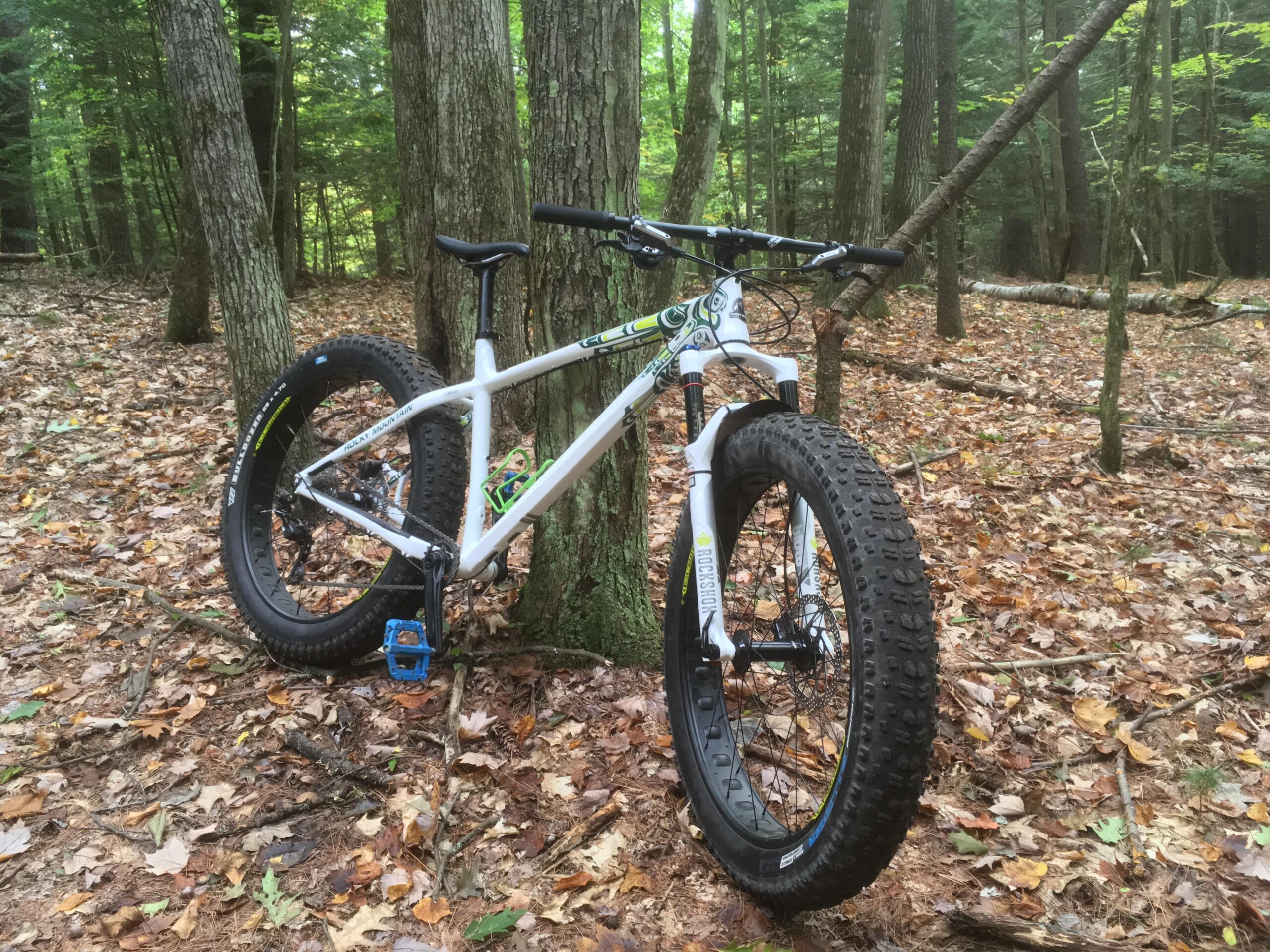 Rocky Mountain Blizzard: A white mountain bike with oversized tires is leaning against a tree in a forested area covered with fallen leaves. The background features tall trees and green foliage, creating a natural setting.