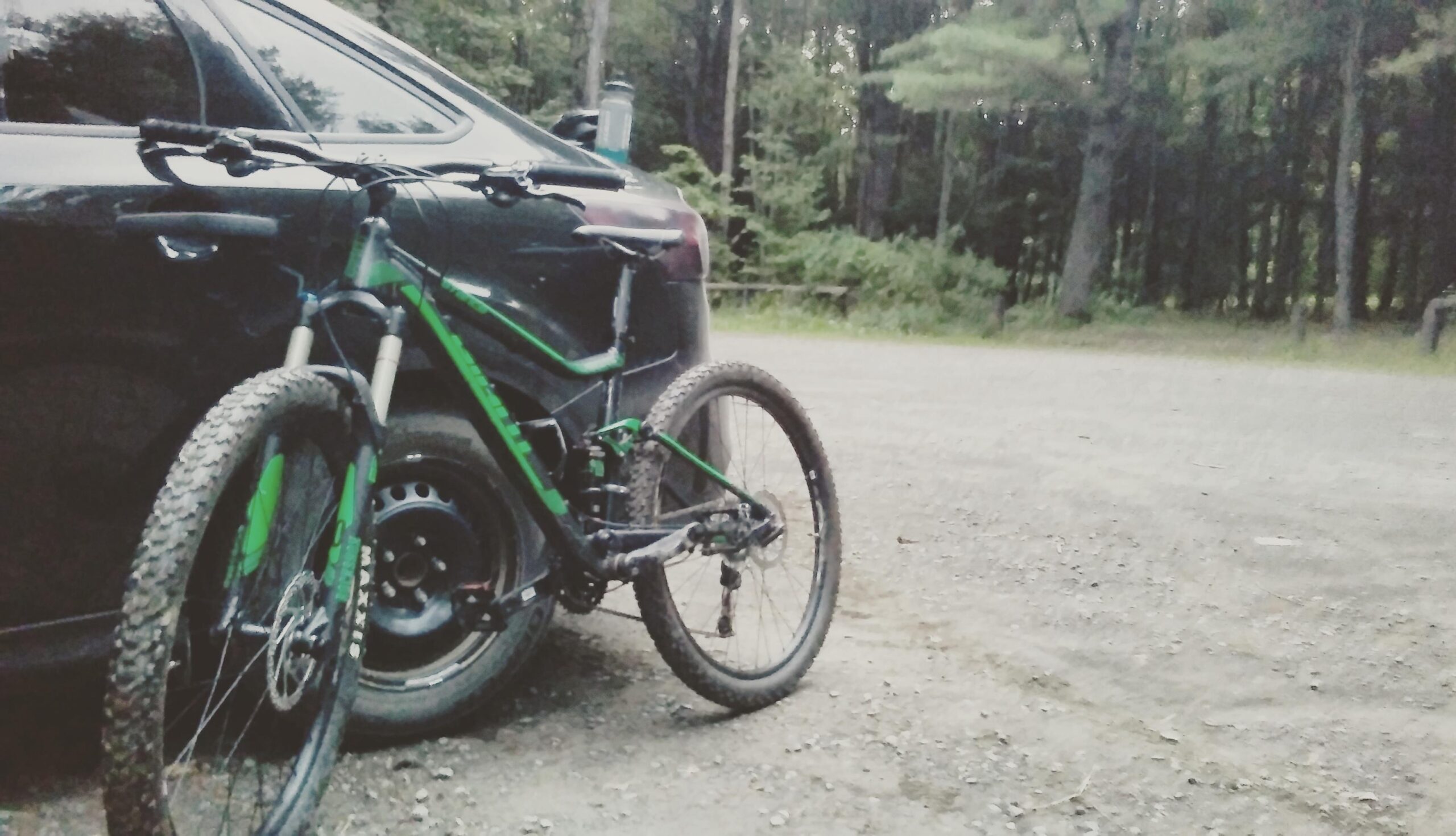 Giant Stance 27.5 2: A mountain bike with green accents is parked next to a black car on a gravel surface, surrounded by trees in a wooded area. The scene suggests an outdoor adventure or cycling activity.