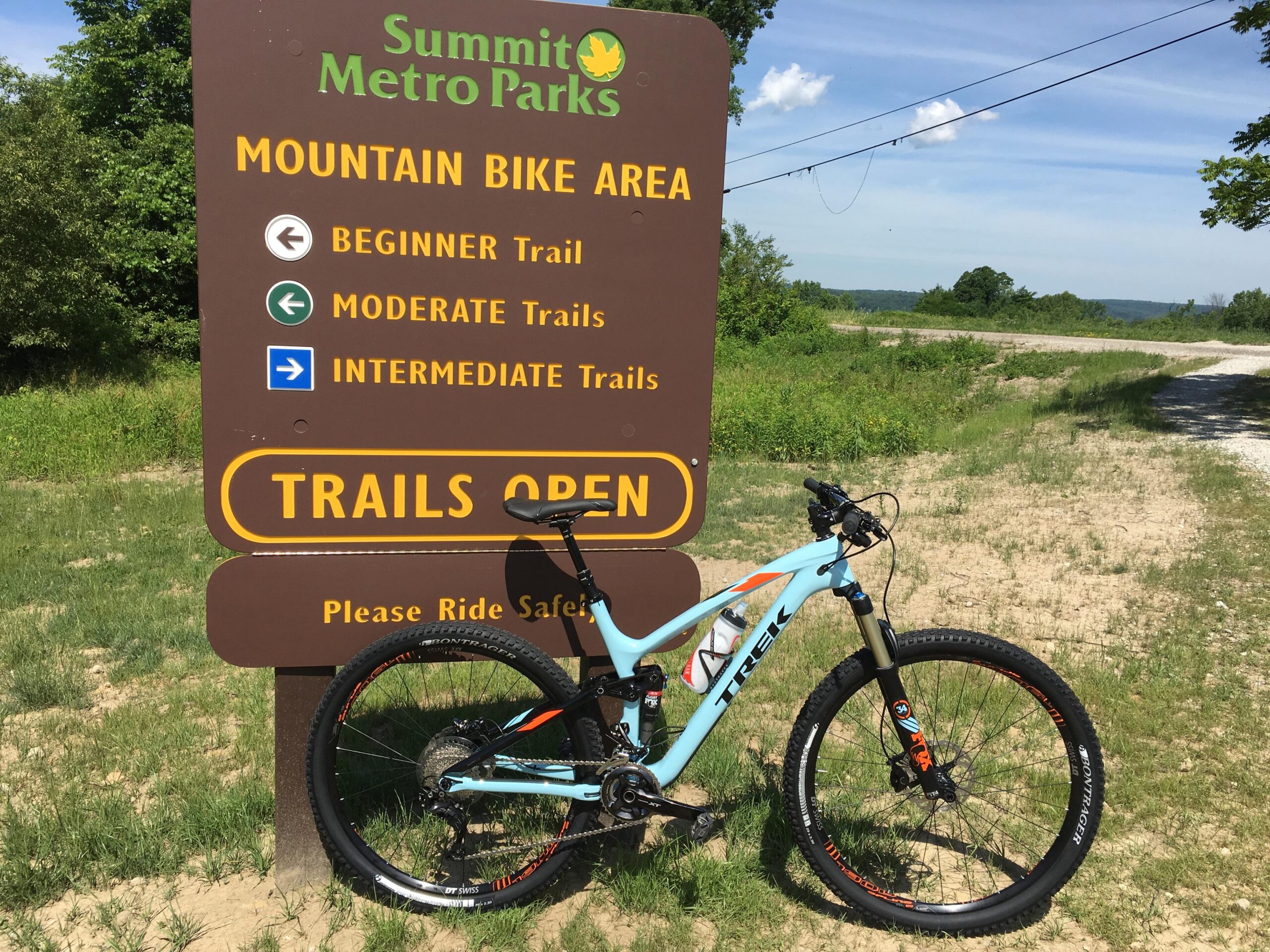 Trek Fuel EX 9.8: A mountain bike parked next to a sign that reads "Summit Metro Parks Mountain Bike Area," indicating trail options: Beginner, Moderate, and Intermediate, with an open status. The scene is set in a natural green environment under a blue sky.