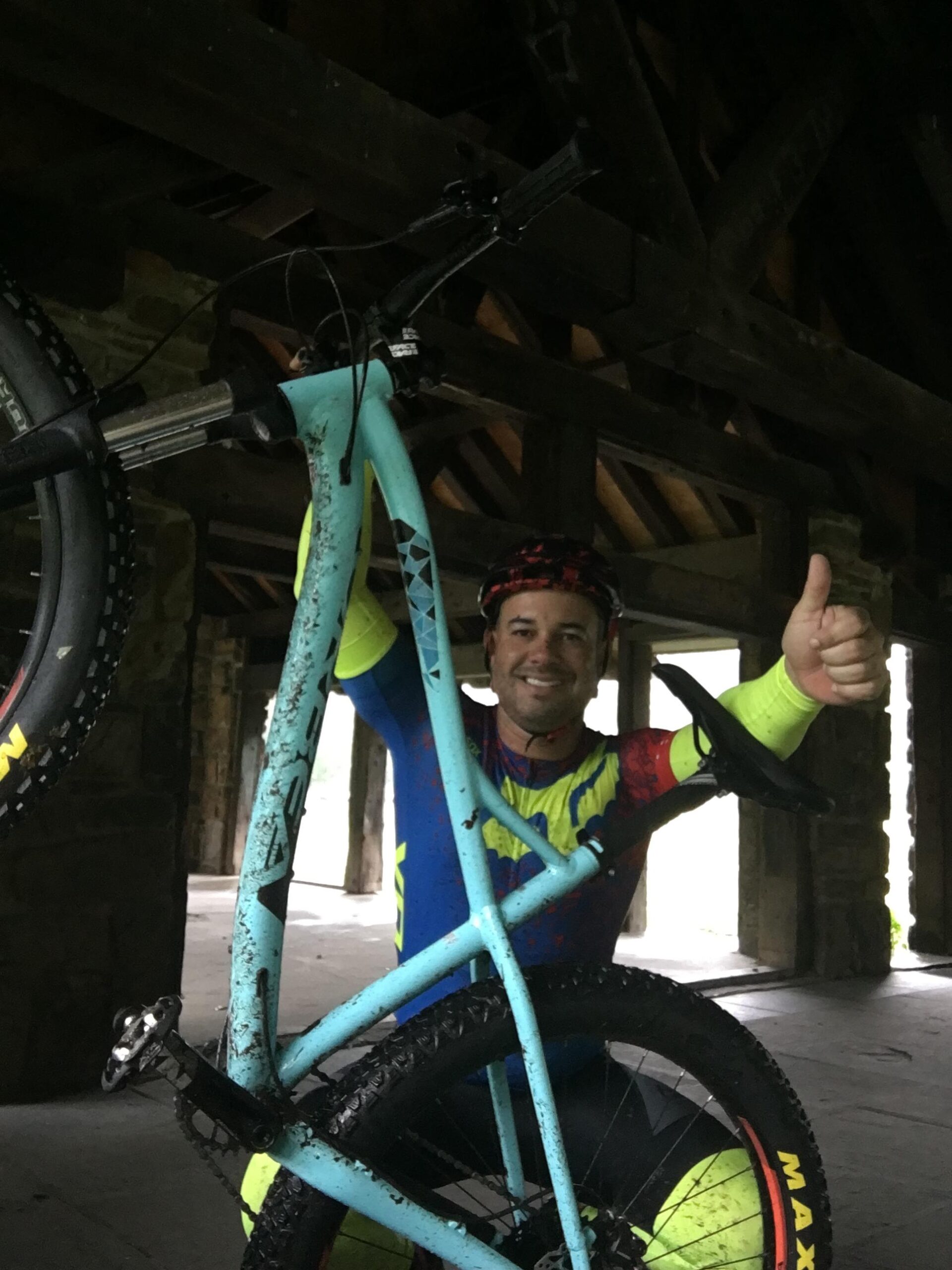 Orbea Loki: A smiling man in a brightly colored cycling outfit holds up a blue mountain bike, giving a thumbs up. The background features a rustic wooden shelter. The bike is slightly muddy, indicating recent use in outdoor activities.