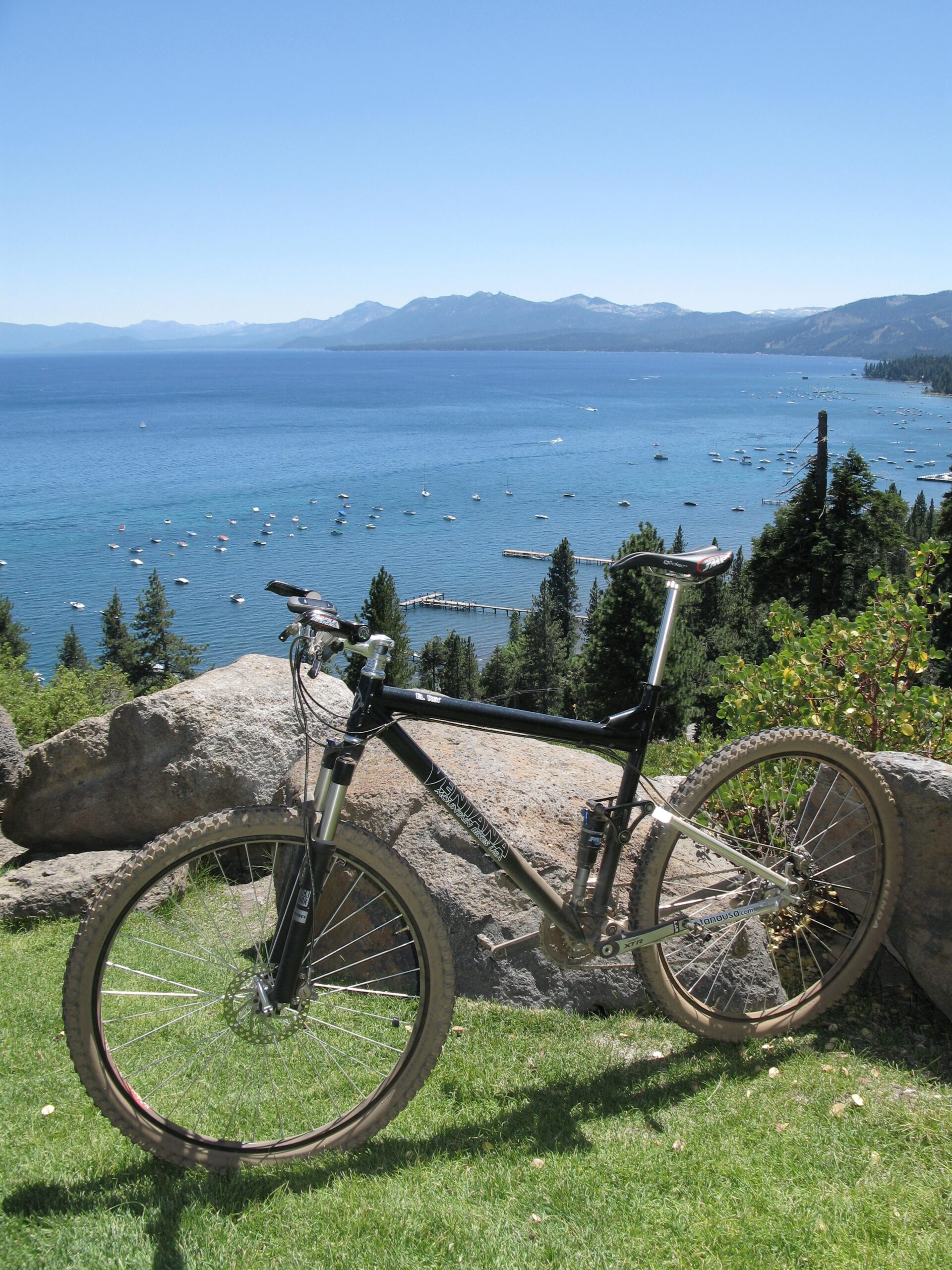 Ventana El Ray El Ray: A mountain bike parked on grassy terrain with large rocks, overlooking a scenic lake surrounded by mountains under a clear blue sky. Various boats are visible on the water, enhancing the outdoor adventure vibe.