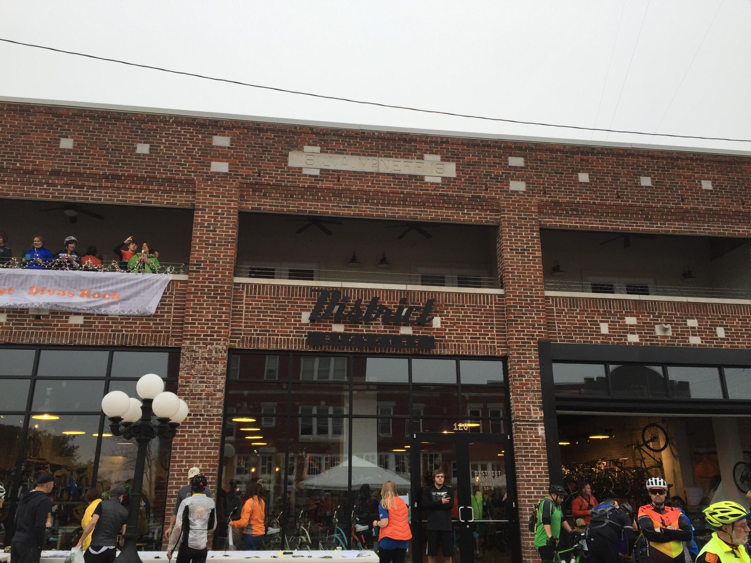 A brick building featuring a large storefront with glass windows. People in colorful attire stand outside, some with bicycles. A decorative banner that reads "Diva's Rock" hangs from the balcony above, where several individuals are gathered. The scene captures a lively atmosphere, likely during an event related to biking.