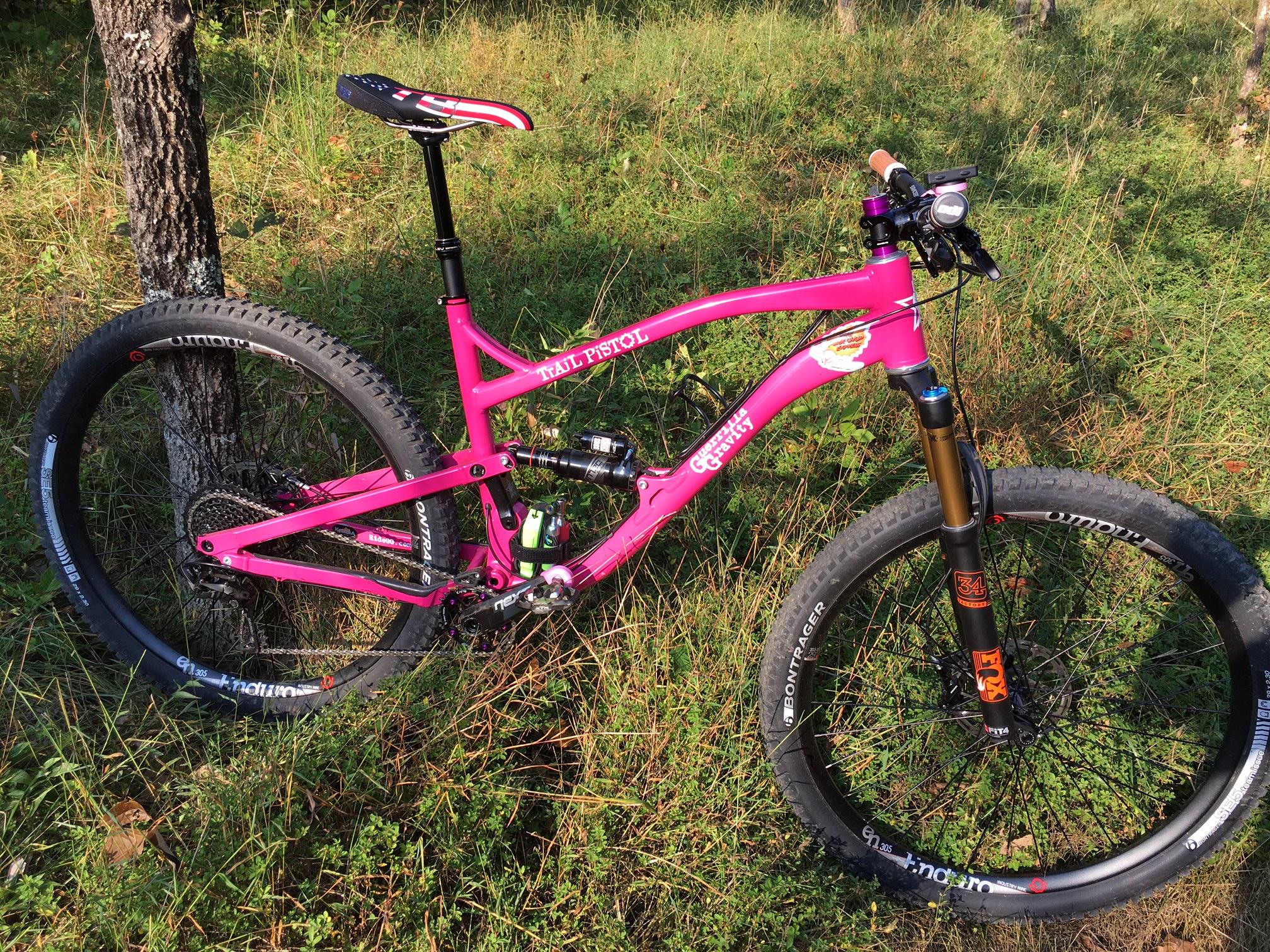 Guerrilla Gravity Trail Pistol: A vibrant pink mountain bike leaning against a tree, set against a backdrop of green grass and foliage. The bike features a suspension fork, wide tires, and a sleek frame, showcasing its design for off-road riding.