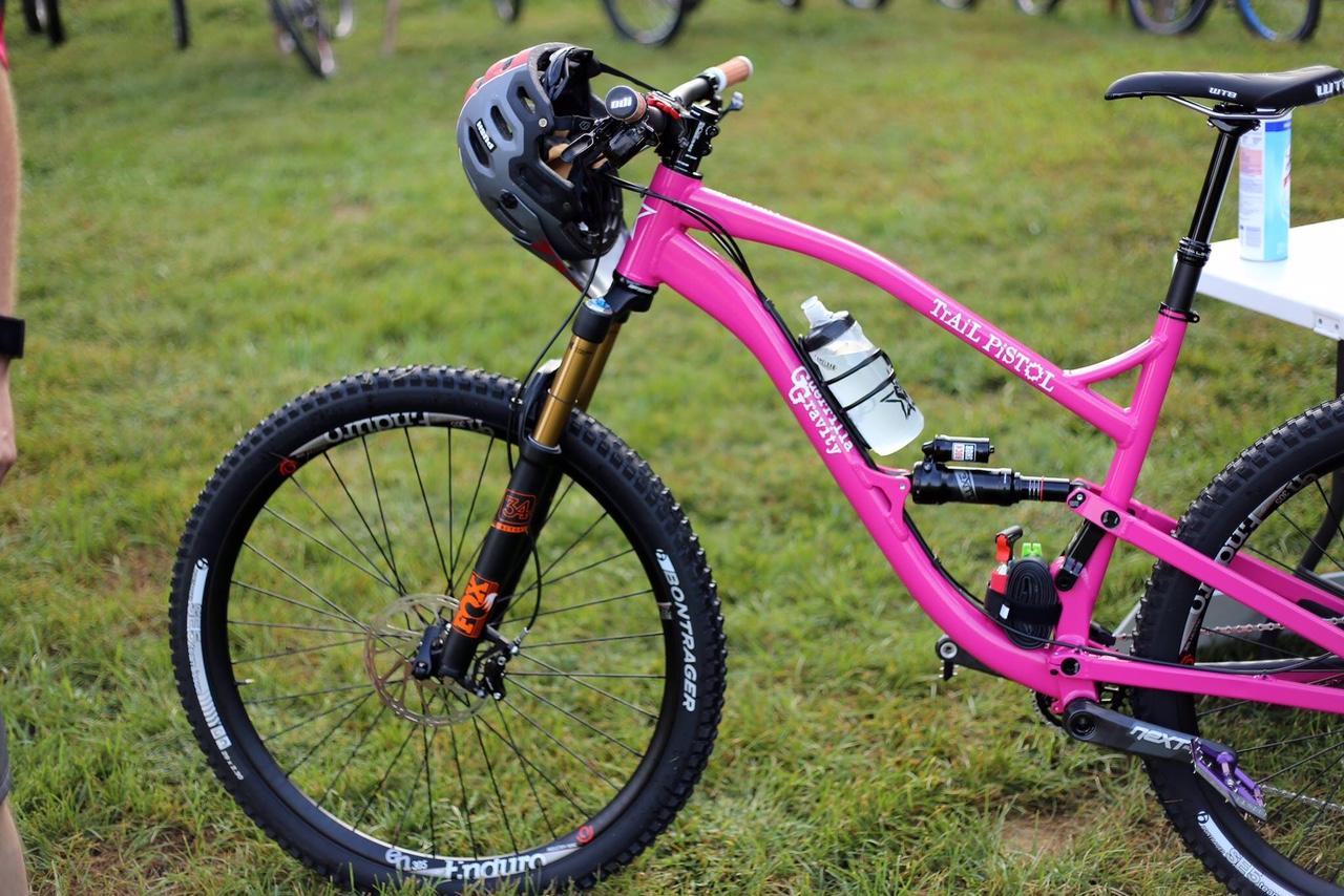 Guerrilla Gravity Trail Pistol: Alt text: A vibrant pink mountain bike with a helmet attached to the handlebars, featuring a water bottle holder, front suspension fork, and rugged tires, resting on green grass.