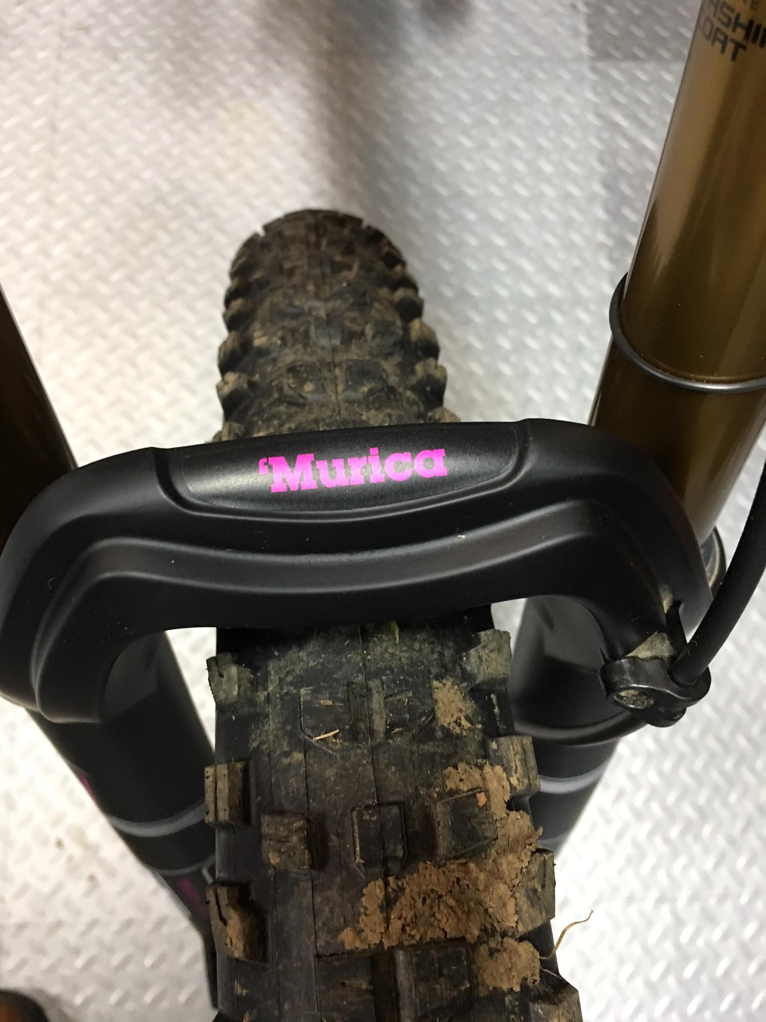Guerrilla Gravity Trail Pistol: A close-up view of a mountain bike's front fork and tire, featuring the word "'Murica" in pink lettering on the fork. The tire shows signs of mud, indicating recent outdoor use, and the background has a textured, light-colored surface.