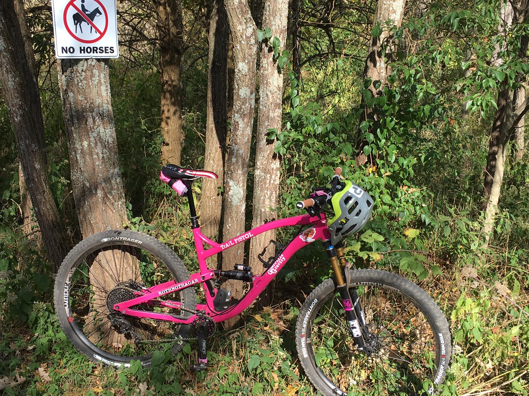 Guerrilla Gravity Trail Pistol: A bright pink mountain bike is leaning against trees in a wooded area, with a sign nearby that reads "No Horses." The bike features a helmet attached and is surrounded by green foliage, indicating a natural setting for outdoor activities.