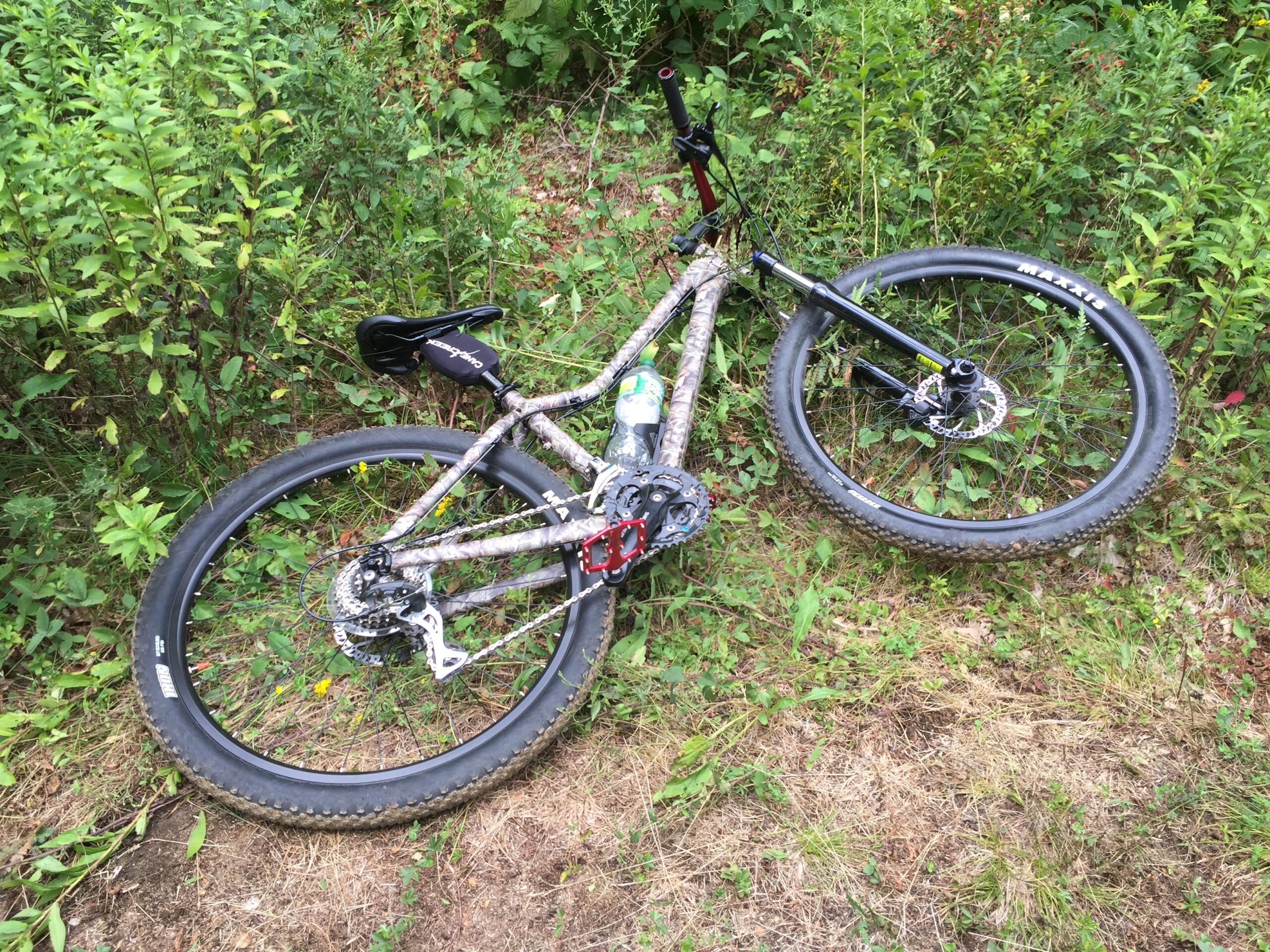 Giant Tempt 4: A mountain bike lies on the ground among tall grass and shrubs, featuring a camouflage-patterned frame. The front wheel is slightly turned, and a water bottle is visible attached to the bike. The setting appears to be a natural outdoor trail or area.