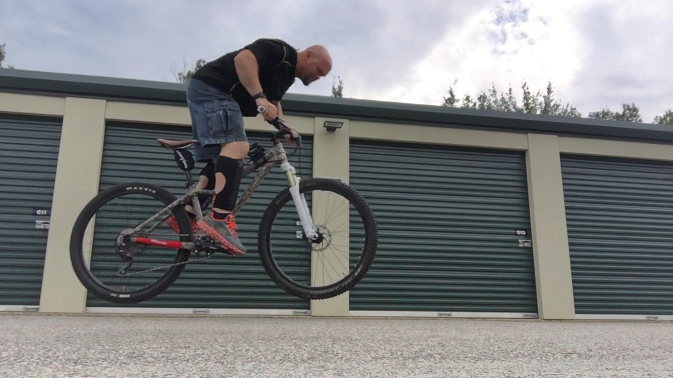 Giant Tempt 4: A person performing a bicycle stunt, lifting the front wheel off the ground while riding a mountain bike in a parking area with storage units in the background. The cyclist is wearing a black shirt, denim shorts, and knee-high socks.