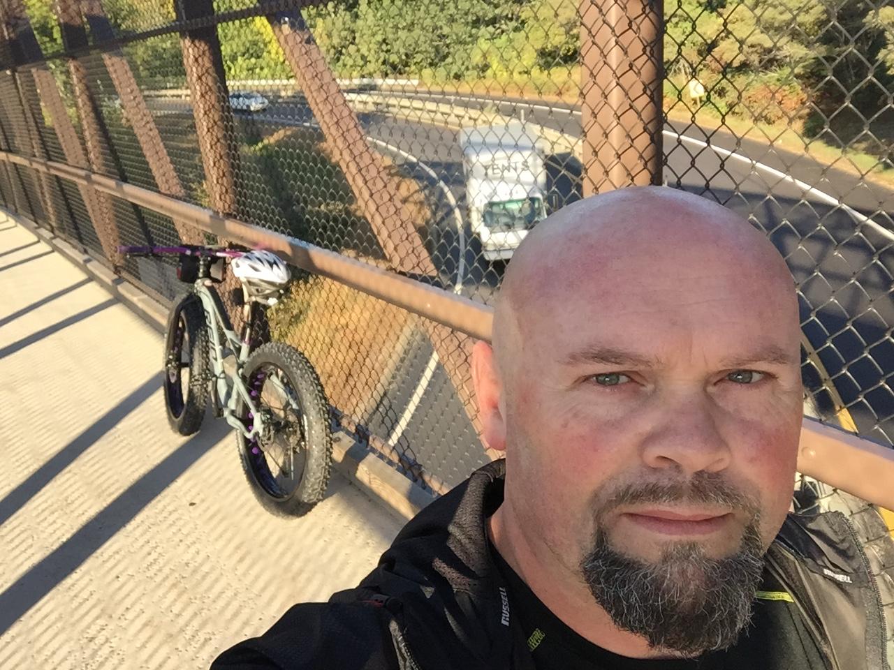 Giant Tempt 4: A person with a bald head and beard stands on a bridge, taking a selfie. A fat tire bike is leaned against the railing beside them. In the background, a winding road can be seen with vehicles passing by and trees lining the edges. The setting is bright and sunny.