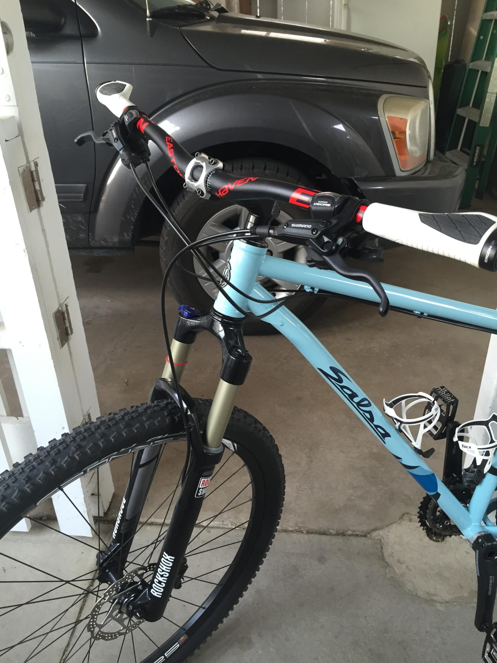 Salsa El Mariachi 3: Alt text: A close-up view of a blue Salsa mountain bike in a garage, featuring black RockShox front forks, a Shimano gear shifter, and white handlebar grips. In the background, a gray vehicle is partially visible.