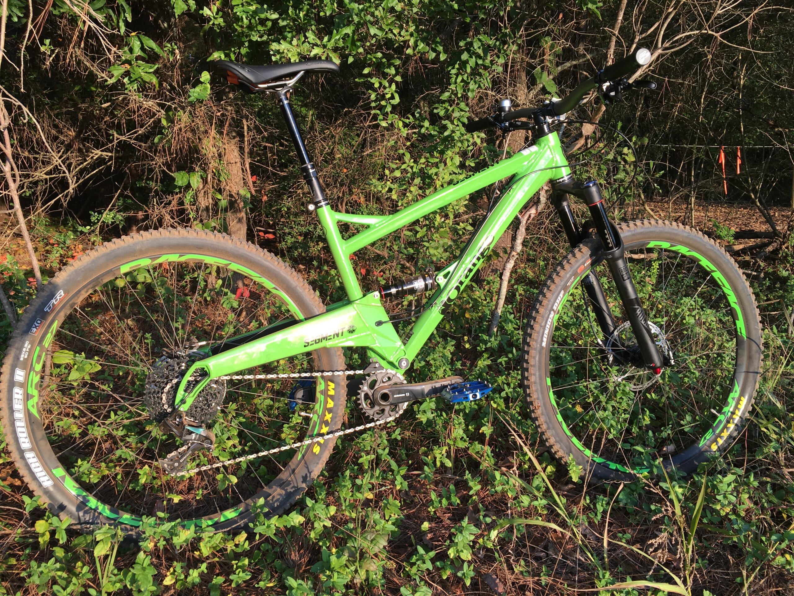 Orange Segment RS: A green mountain bike with a black front fork, positioned near a dense backdrop of foliage and shrubs. The bike features thick, rugged tires and is resting on a patch of grass. Sunlight highlights the vibrant colors of the bike and surrounding vegetation.