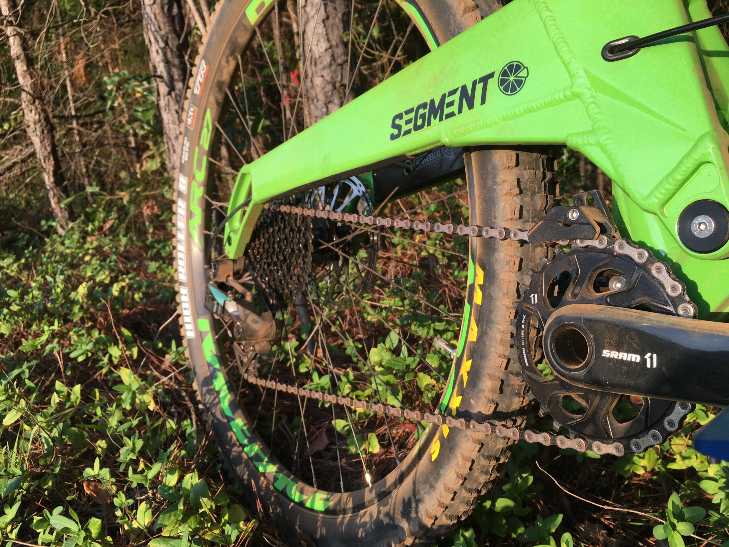 Orange Segment RS: A close-up view of a bright green mountain bike frame, showcasing the rear wheel, chain, and drivetrain components. The bike is set against a backdrop of trees and foliage, with sunlight illuminating the scene. The image highlights details such as the "SEGMENT" logo on the frame, the SRAM crankset, and the tire tread pattern.