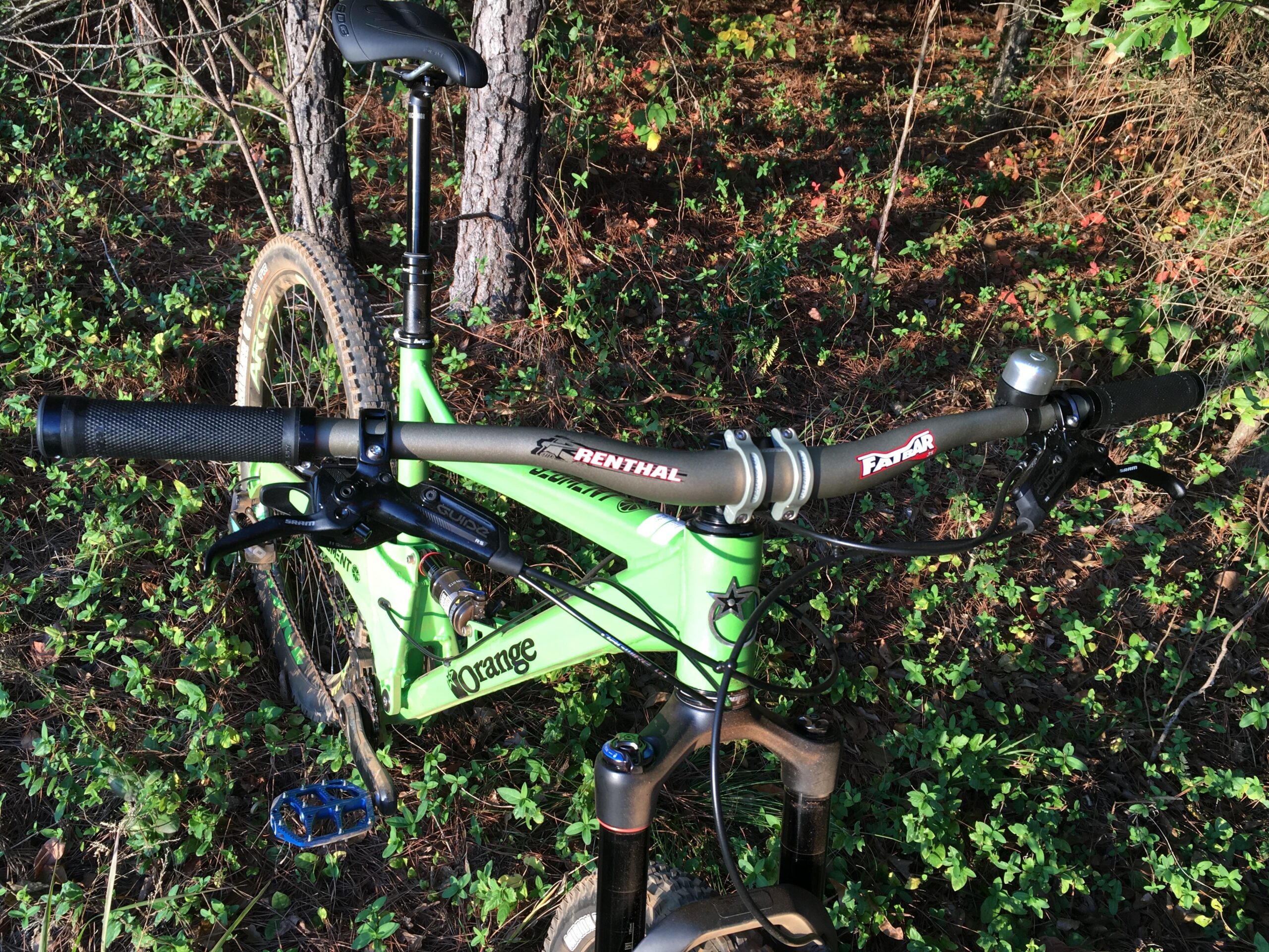 Orange Segment RS: A close-up view of a green mountain bike standing on the ground among foliage. The image focuses on the handlebars, grips, and brake levers, showcasing details such as the Renthal branding on the handlebars, and the Orange logo on the frame. The background features trees and greenery, indicating a natural outdoor setting.