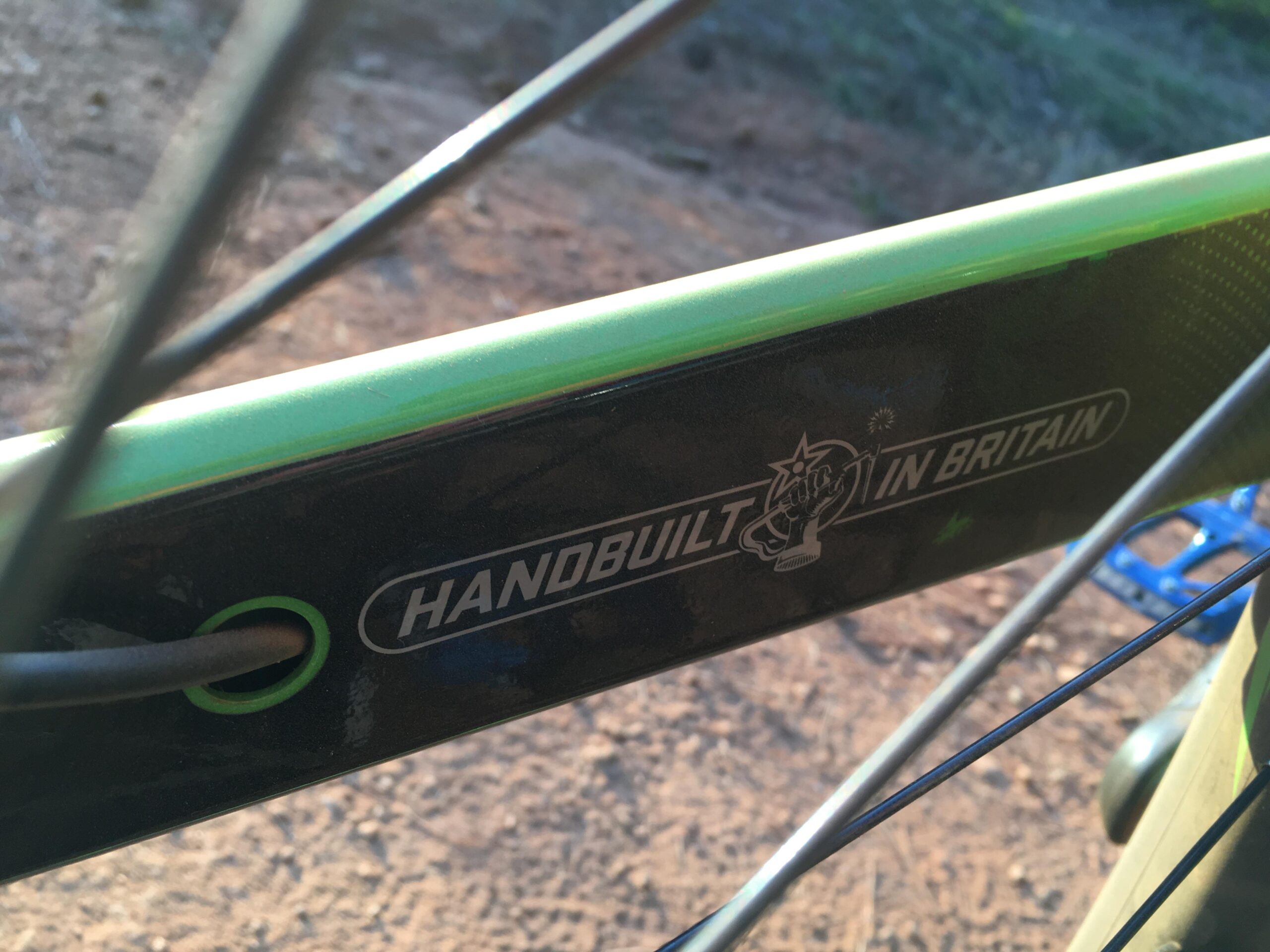 Orange Segment RS: Close-up view of a bicycle frame displaying the text "HANDBUILT IN BRITAIN." The frame features a sleek design with a green accent. The background shows a dirt surface, suggesting an outdoor setting, with part of the bicycle's wheel and components visible.