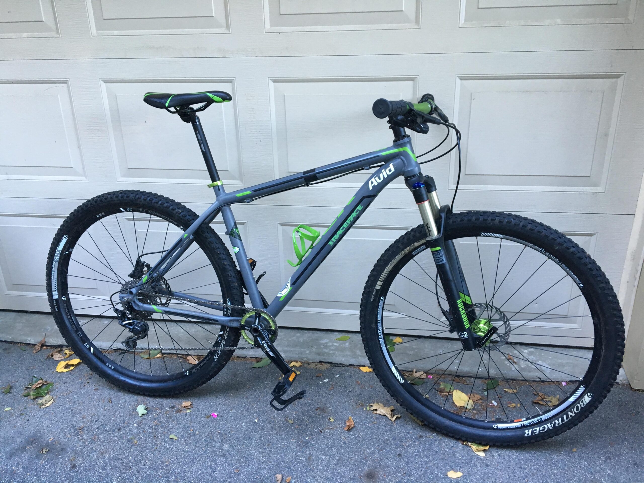 Trek Rig: A gray mountain bike with green accents, featuring a lightweight frame, thick tires, and a front suspension fork. The bike is parked against a garage door, with some fallen leaves on the ground nearby.