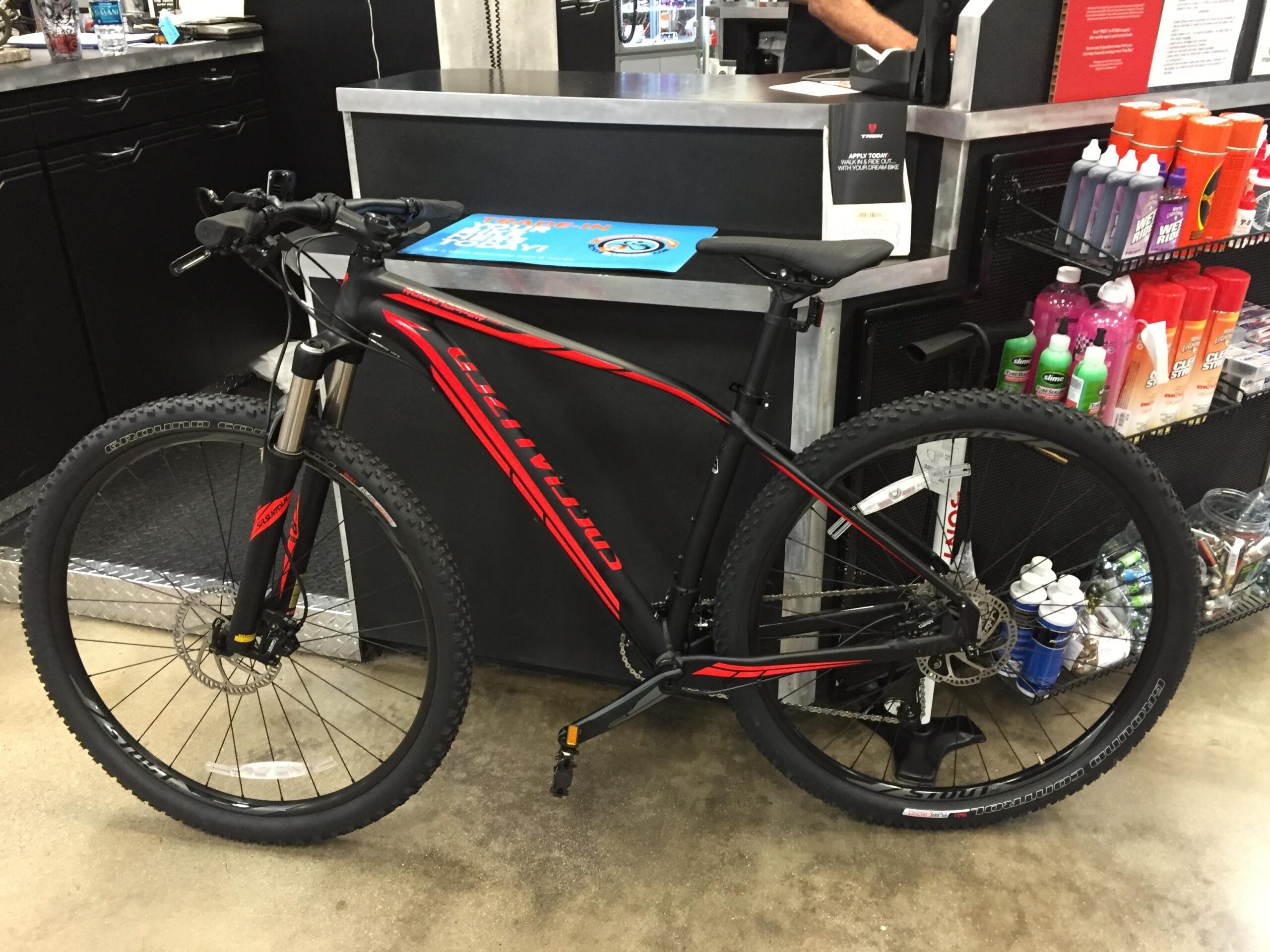 Specialized Rockhopper 29: A black and red mountain bike is leaning against a counter in a store. The bike features thick tires, front suspension, and a sleek design. In the background, various products are displayed on the counter, including bottles of cleaning supplies and promotional materials.