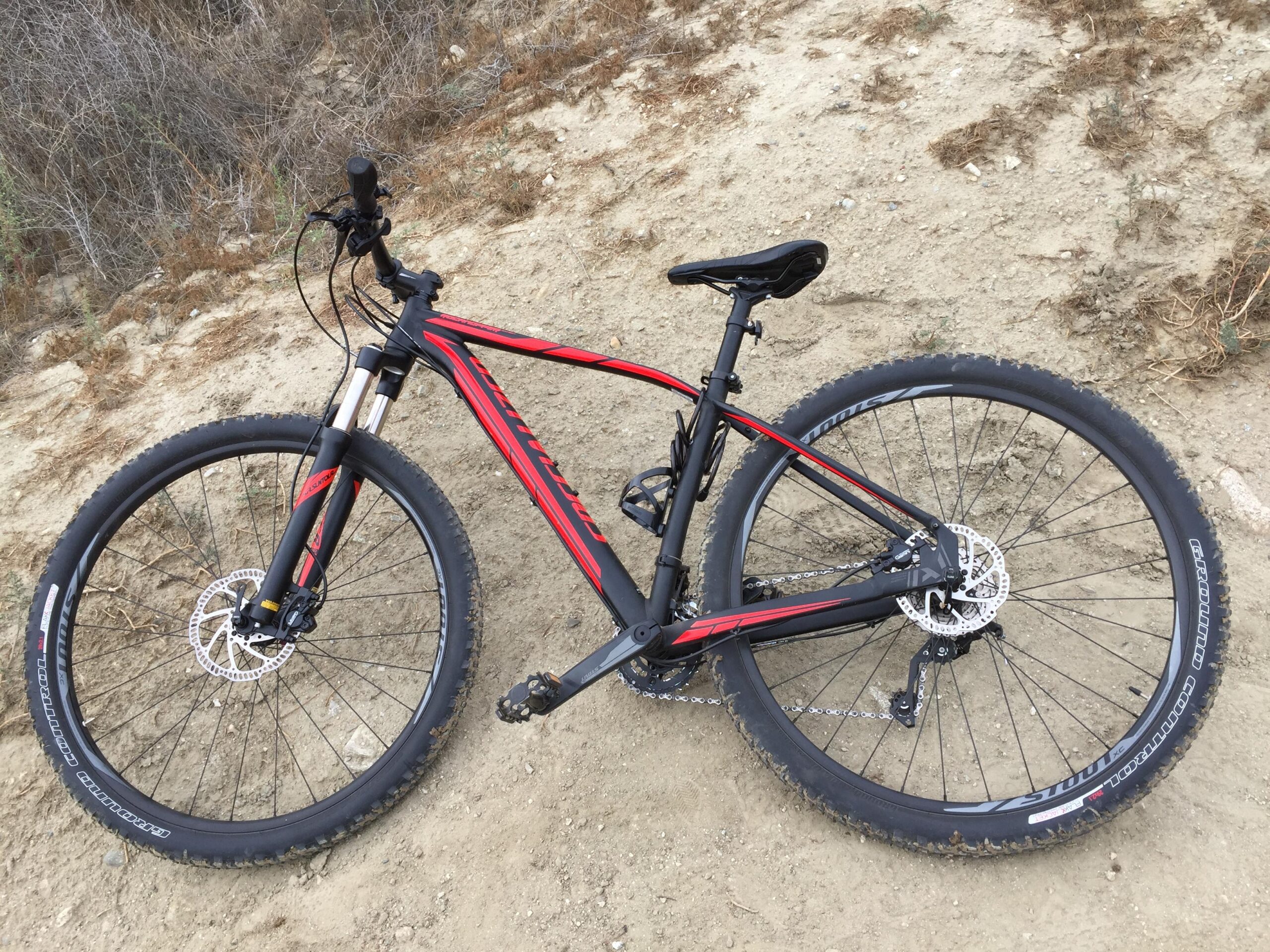 Specialized Rockhopper 29: A black and red mountain bike lying on a dirt path, with its front wheel angled to the side and the drivetrain visible. Surrounding vegetation is sparse, with dry grass and soil visible in the background.