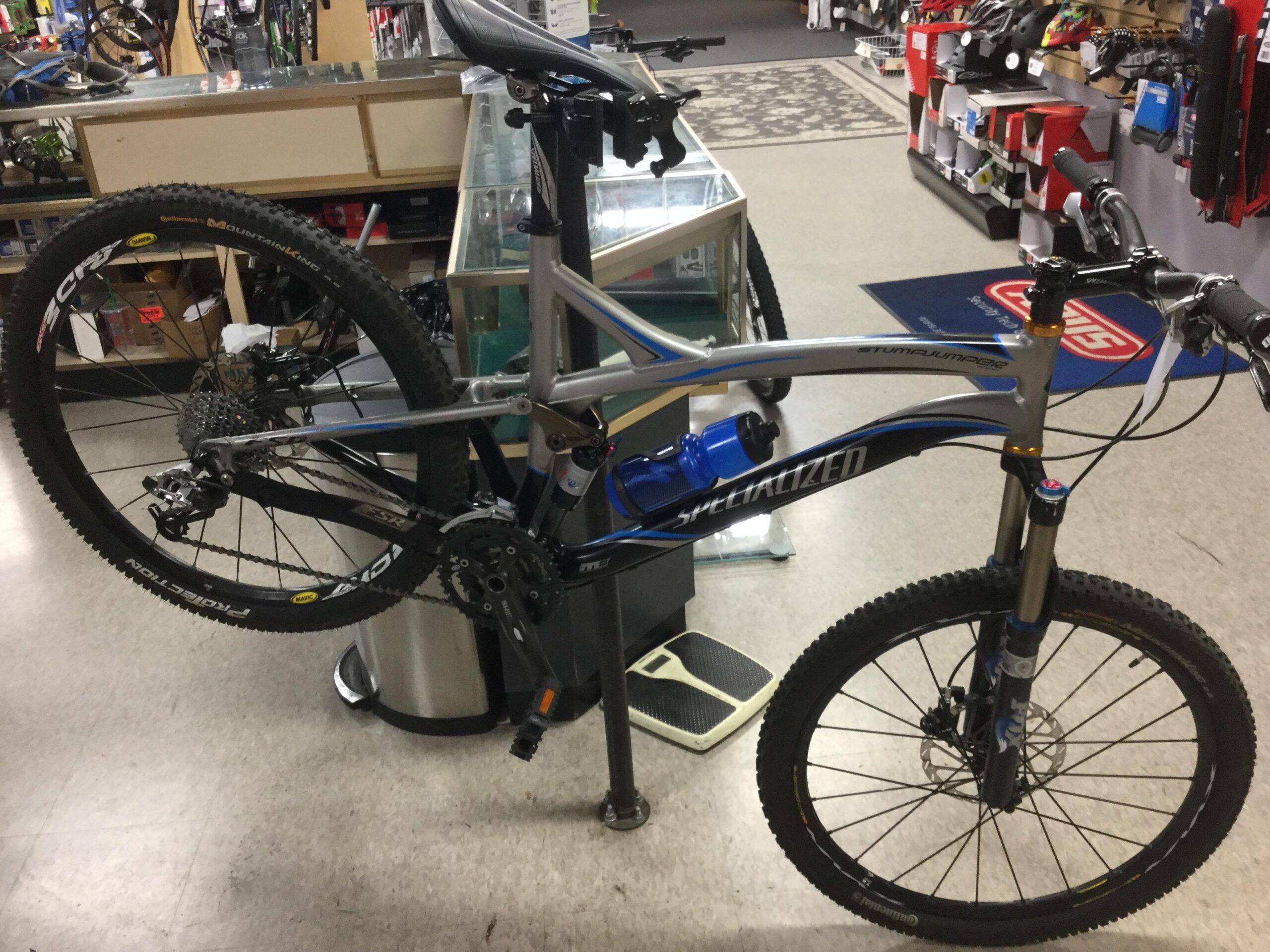 Specialized Stumpjumper FSR: A mountain bike displayed in a retail store, featuring a silver and blue frame, wide tires with tread for traction, and a visible suspension system. The bike is positioned on a stand, showcasing its components and features. In the background, various bicycle accessories and tools are visible.