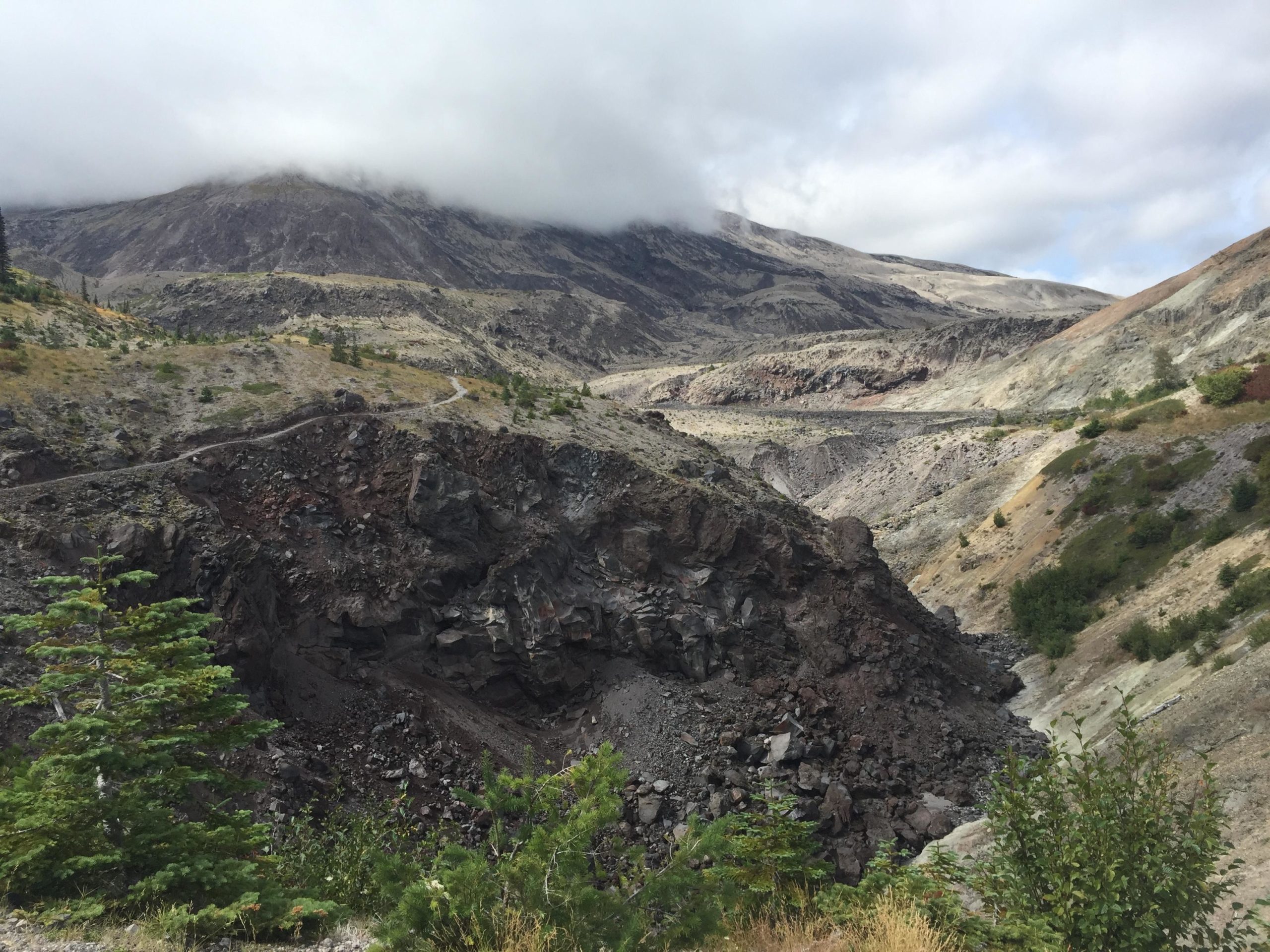 A rugged mountainous landscape with rocky slopes and a valley in the foreground. The terrain shows signs of volcanic activity, with dark, rocky outcrops and scattered greenery. Clouds partially obscure the peaks, creating a dramatic sky. A winding path is visible on the hillside, indicating a trail for hikers. Ape Canyon#234, Abraham#216d, Smith Creek#225 Trails mountain bike trail.