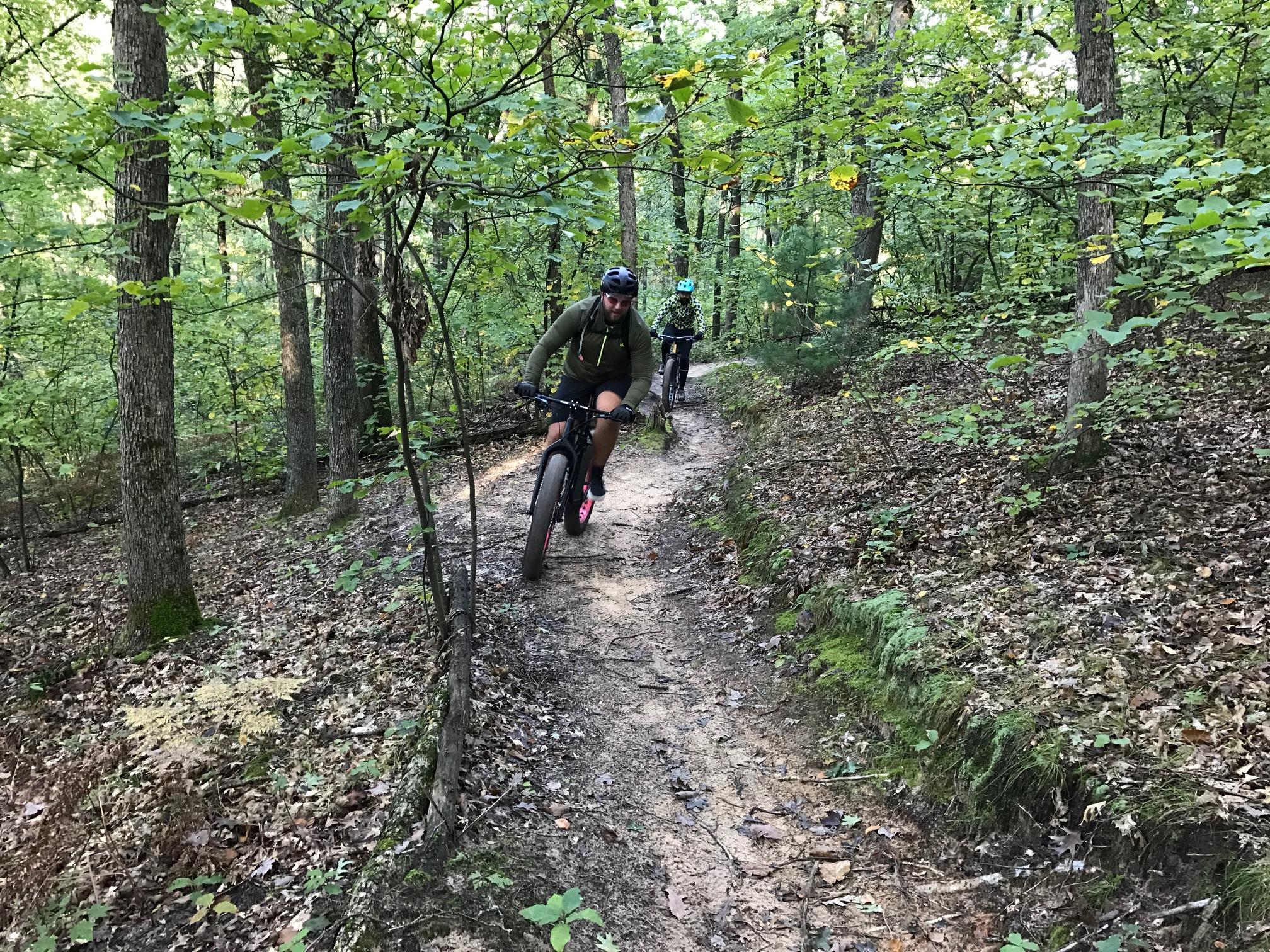 Two cyclists navigate a winding dirt trail through a lush green forest. The first cyclist, wearing a green hoodie and a helmet, is in the foreground, while a second cyclist in a blue helmet is visible in the background. The scene is surrounded by trees and abundant foliage, showcasing the peacefulness of nature during daytime. Levis Mounds mountain bike trail.