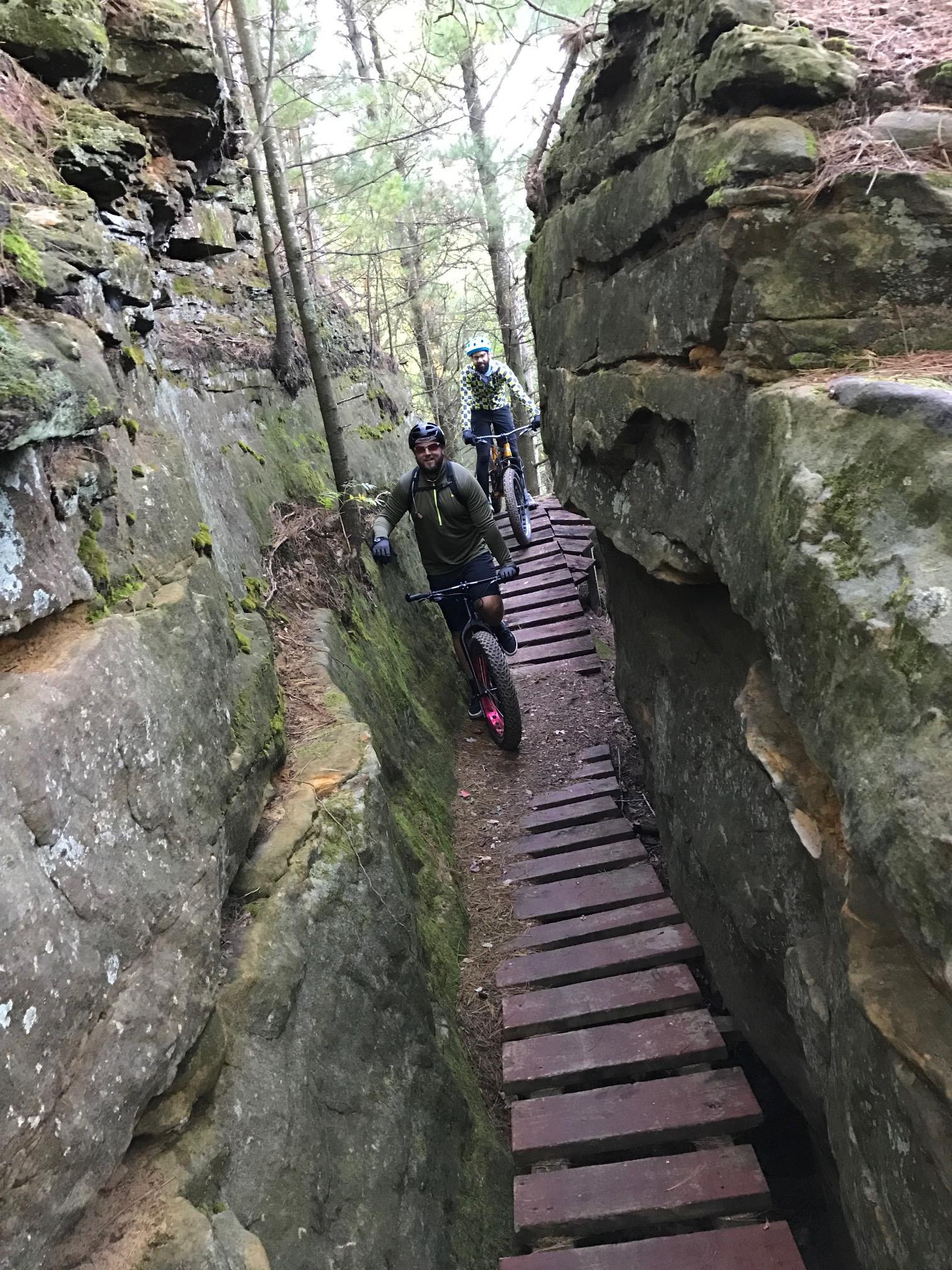 Mountain bikers navigating a narrow trail surrounded by rocky cliffs and trees. Wooden planks form a bridge over the uneven ground, showcasing an adventurous outdoor setting. Levis Mounds mountain bike trail.