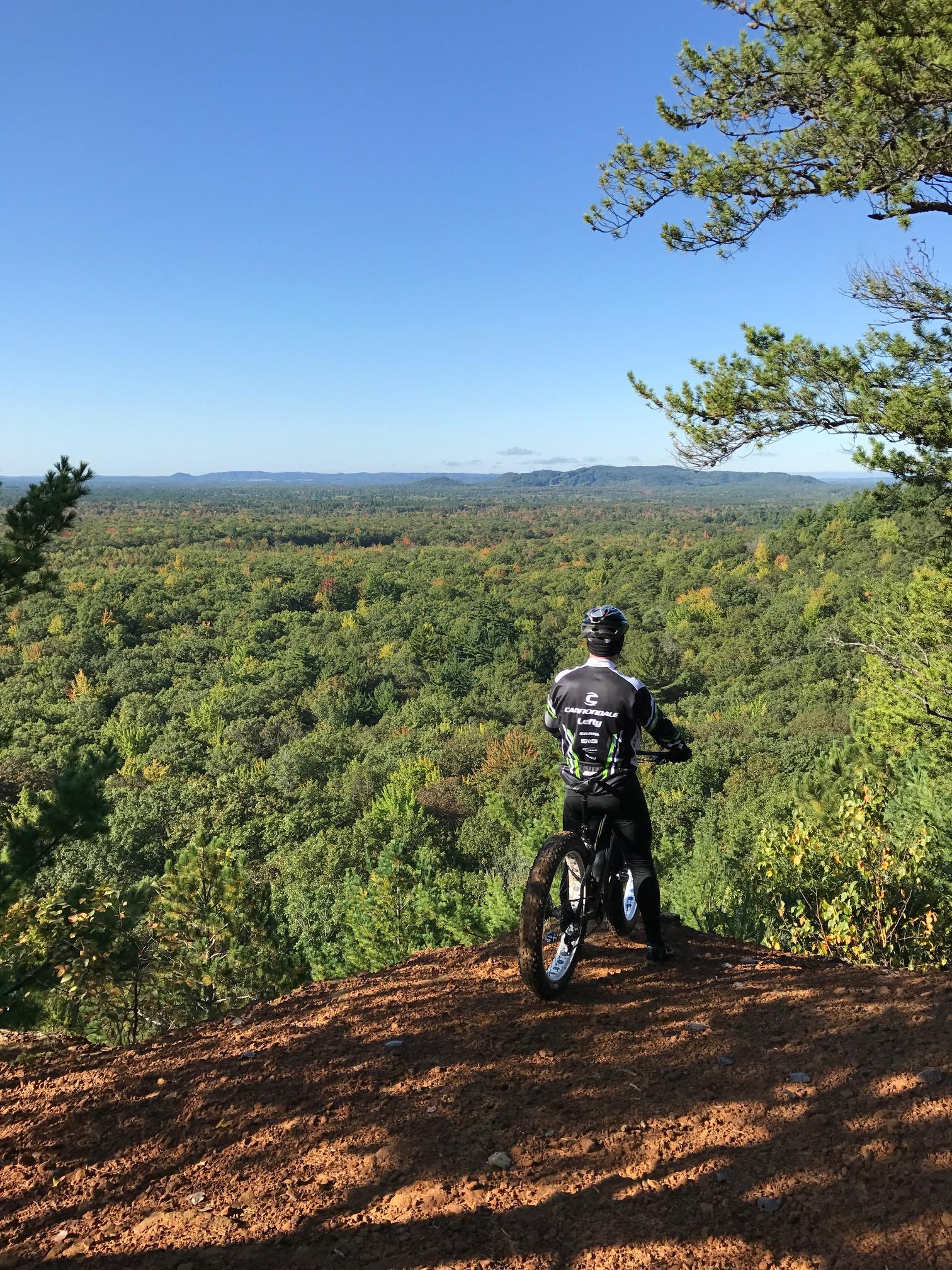 A cyclist in a black and white jersey stands on a dirt cliff, overlooking a vast expanse of lush green forest and distant hills under a clear blue sky. The scene captures the serenity of nature and the thrill of outdoor adventure. Levis Mounds mountain bike trail.