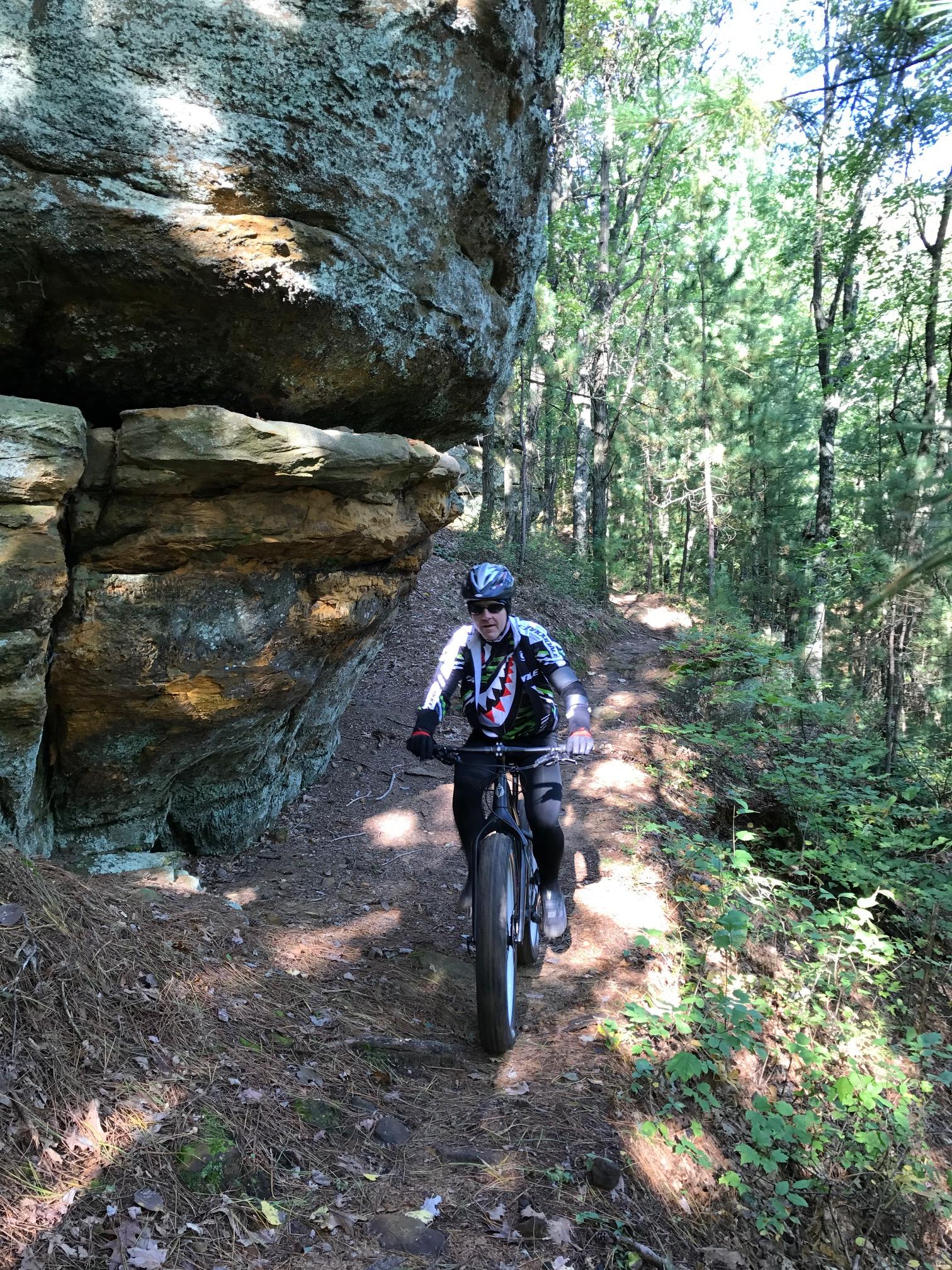 A mountain biker riding on a narrow trail surrounded by trees, with a large rock formation to the left. The cyclist is wearing a helmet and a jersey with a shark design, navigating through the forested area. Levis Mounds mountain bike trail.