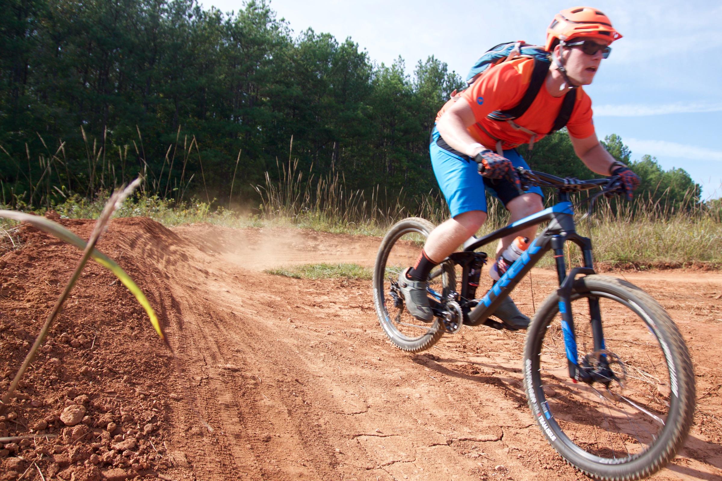 A mountain biker riding on a dirt trail, kicking up dust as they navigate a turn. The cyclist is wearing an orange helmet and shirt, blue shorts, and protective gear, with a backpack visible. The background features green trees and tall grass, indicating a natural outdoor setting. USNWC mountain bike trail.