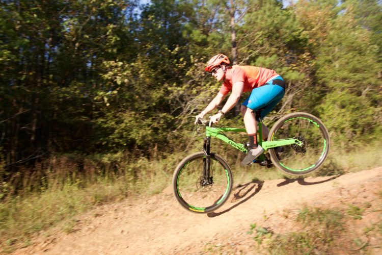 A cyclist in an orange shirt and blue shorts riding a green mountain bike down a dirt path surrounded by trees. The bike is airborne as it goes over a small hill, capturing a moment of action and adventure in nature.