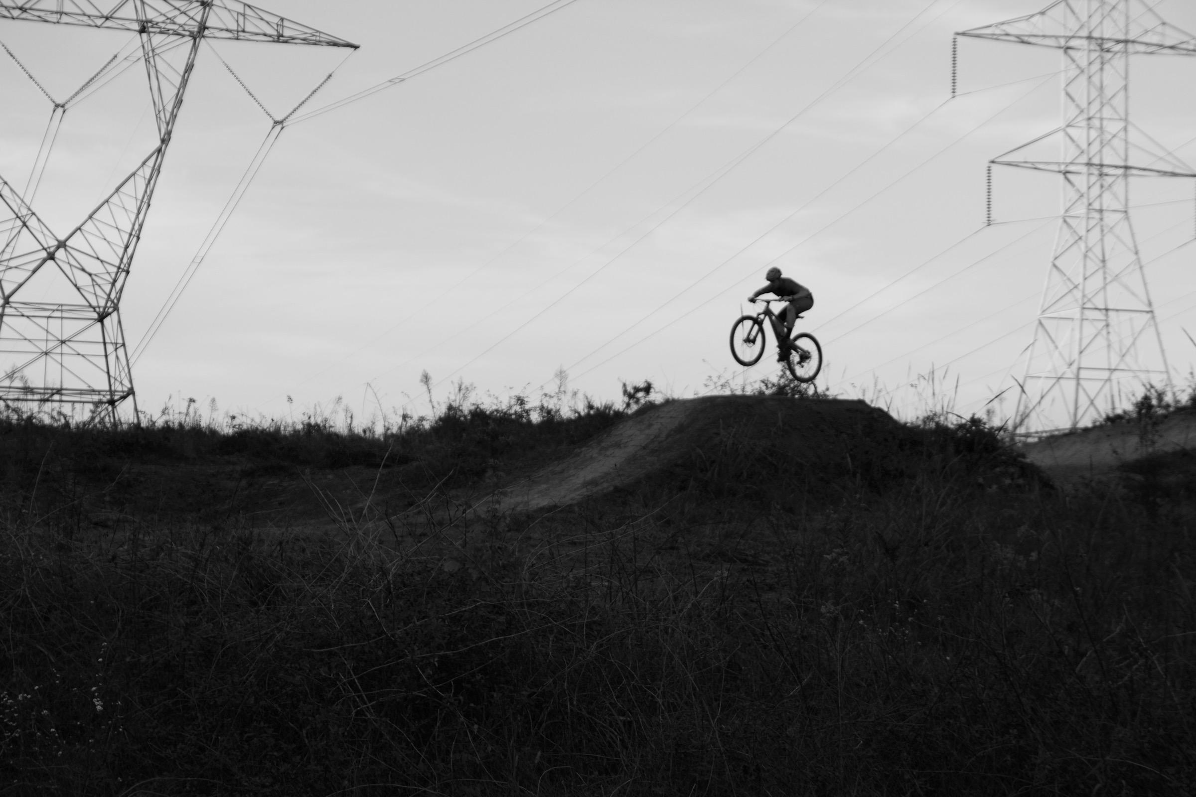 A silhouette of a cyclist performing a jump on a dirt ramp, with tall power lines and a cloudy sky in the background. The image is in black and white, emphasizing the action and the landscape. USNWC mountain bike trail.