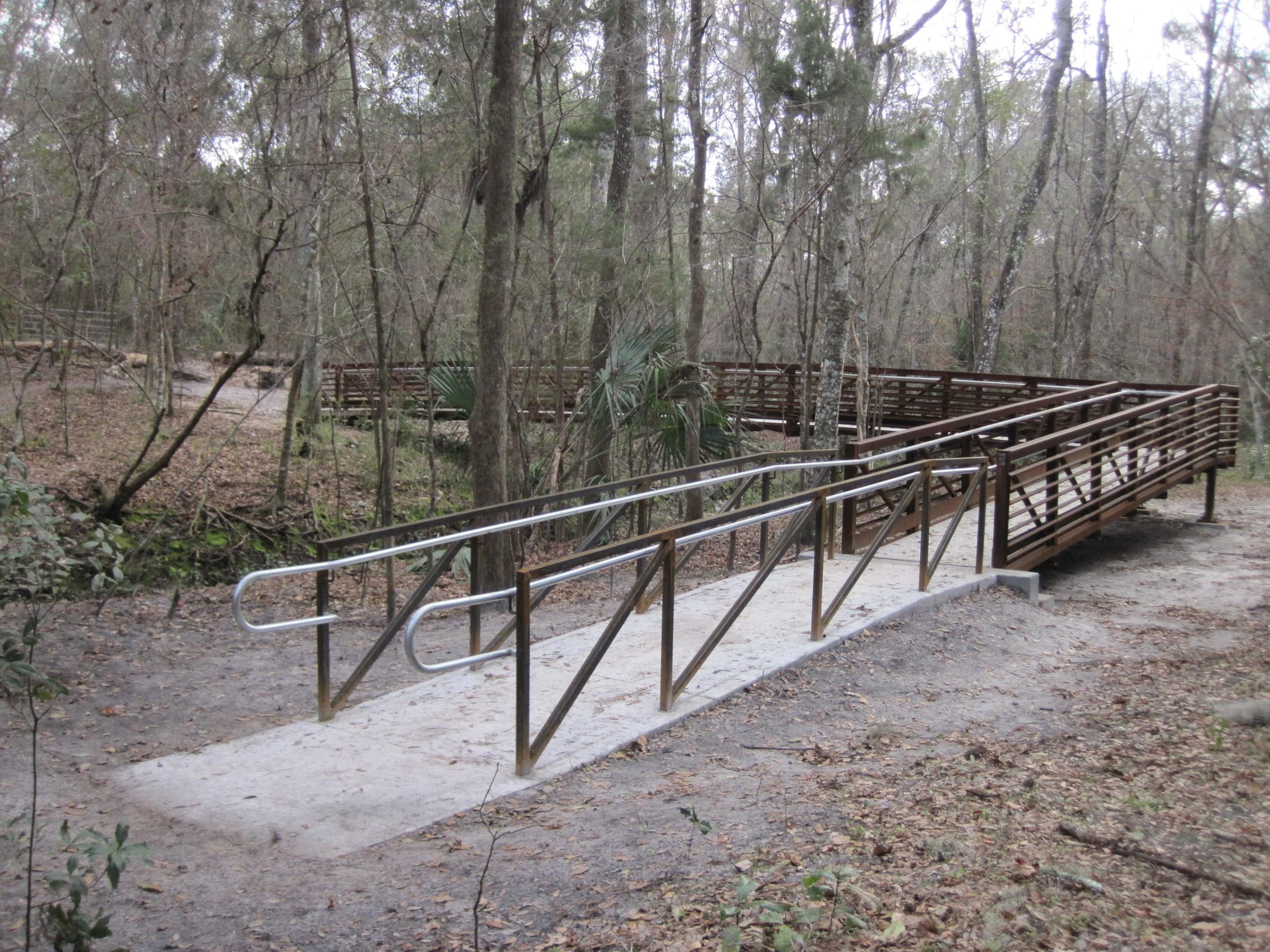 A wooden bridge with a ramp, set in a wooded area. The bridge leads across a small creek and is surrounded by trees with bare branches. The path beside the bridge is covered with fallen leaves and appears to have well-maintained trails. Sweetwater Preserve mountain bike trail.