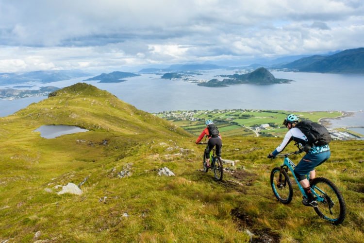 Two mountain bikers navigating a hilly terrain overlooking a scenic view of a fjord and islands. Lush green grass covers the hills, with a small pond visible on the left. The sky is partly cloudy, providing a dramatic backdrop to the landscape.