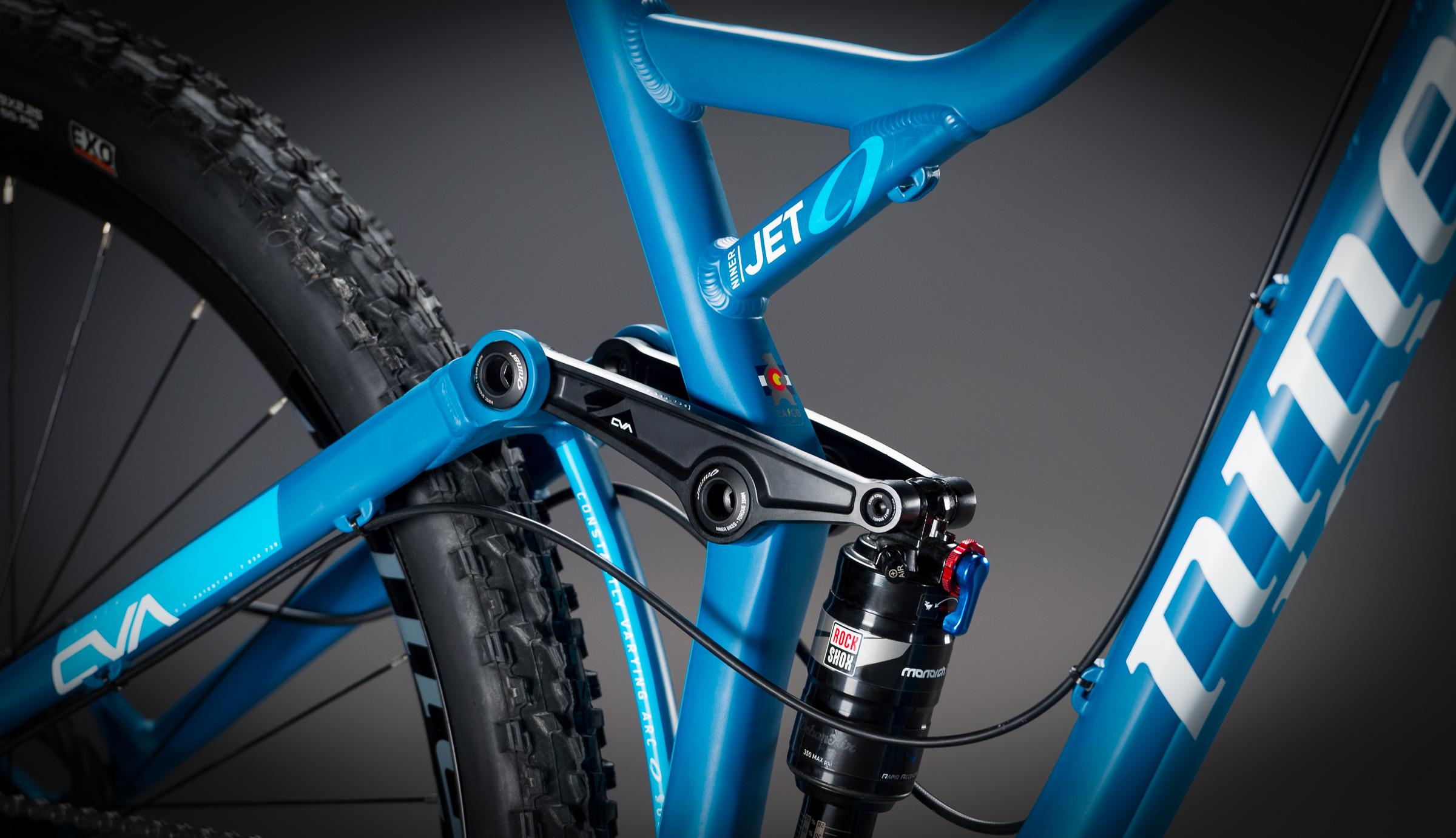 Niner Jet 9: Close-up view of a blue mountain bike frame featuring the suspension system, including the rear shock and linkage. The image showcases the bike's design details, including branding and the intricacies of the frame construction. The background is a smooth gradient, enhancing the focus on the bike components.