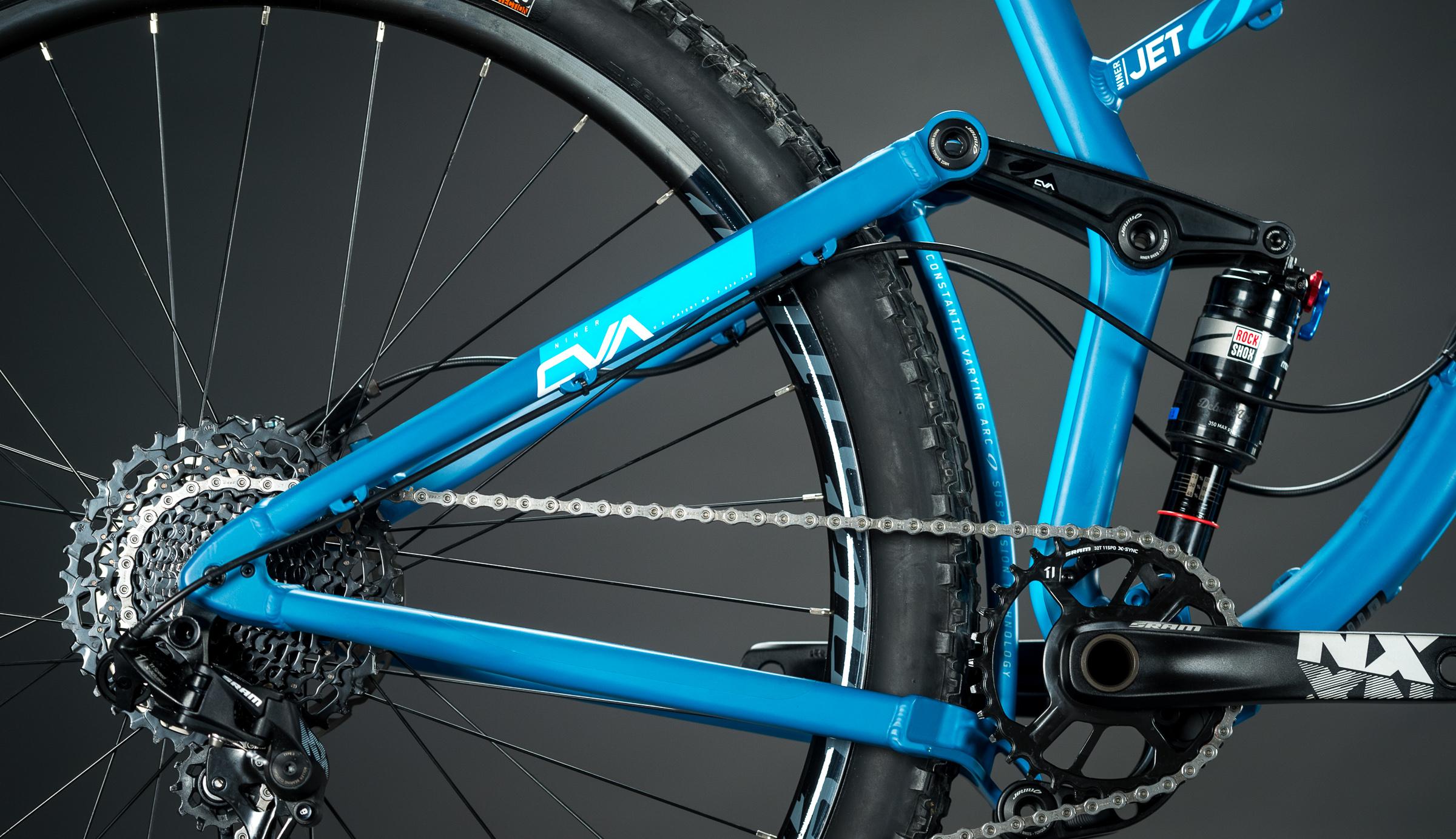 Niner Jet 9: Close-up view of the rear section of a blue mountain bike, highlighting the suspension system, drivetrain, and rear wheel. The image showcases intricate components such as the cassette, chain, and shock absorber against a dark background.