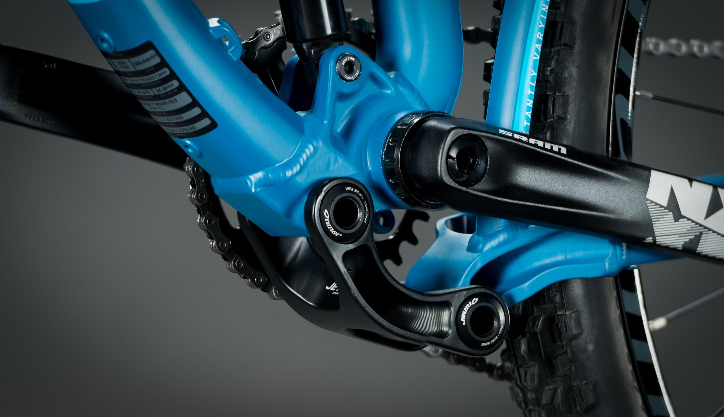 Niner Jet 9: Close-up view of a blue bicycle frame's bottom bracket area, featuring a SRAM crankset and chainring, with visible chain links. The image highlights the intricate design of the bike's components against a dark background.