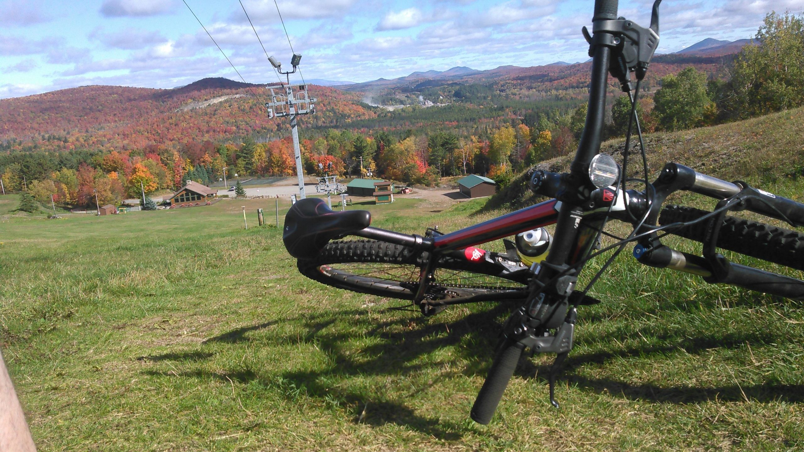 A mountain bike resting on green grass, with a scenic view of colorful autumn trees and mountains in the background. A ski lift is visible nearby, along with a lodge and other buildings in the valley below. The sky is partly cloudy, creating a picturesque outdoor setting. Mount Pisgah Ski Area mountain bike trail.