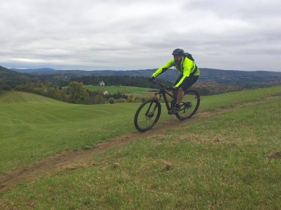 A cyclist wearing a bright yellow jacket rides along a dirt path on a grassy hill, with a panoramic view of rolling hills and a cloudy sky in the background. Kingdom Trails mountain bike trail.