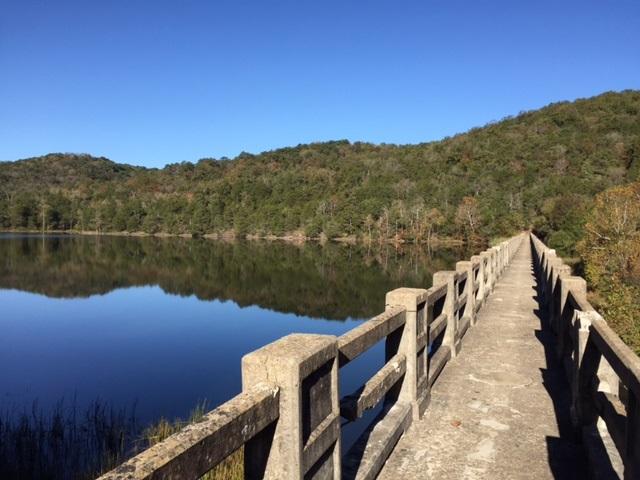 A serene view of a concrete path extending over a calm lake, surrounded by lush green hills under a clear blue sky. The water reflects the landscape, creating a peaceful atmosphere. Lake Leatherwood Gravity Project mountain bike trail.