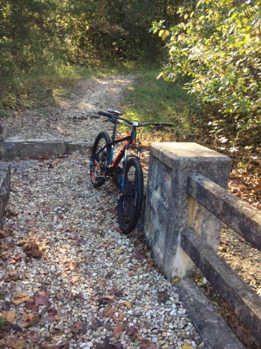 A mountain bike parked beside a stone barrier on a gravel path, surrounded by trees and foliage in a wooded area during autumn. Leaves are scattered on the ground, indicating the season. Lake Leatherwood Gravity Project mountain bike trail.