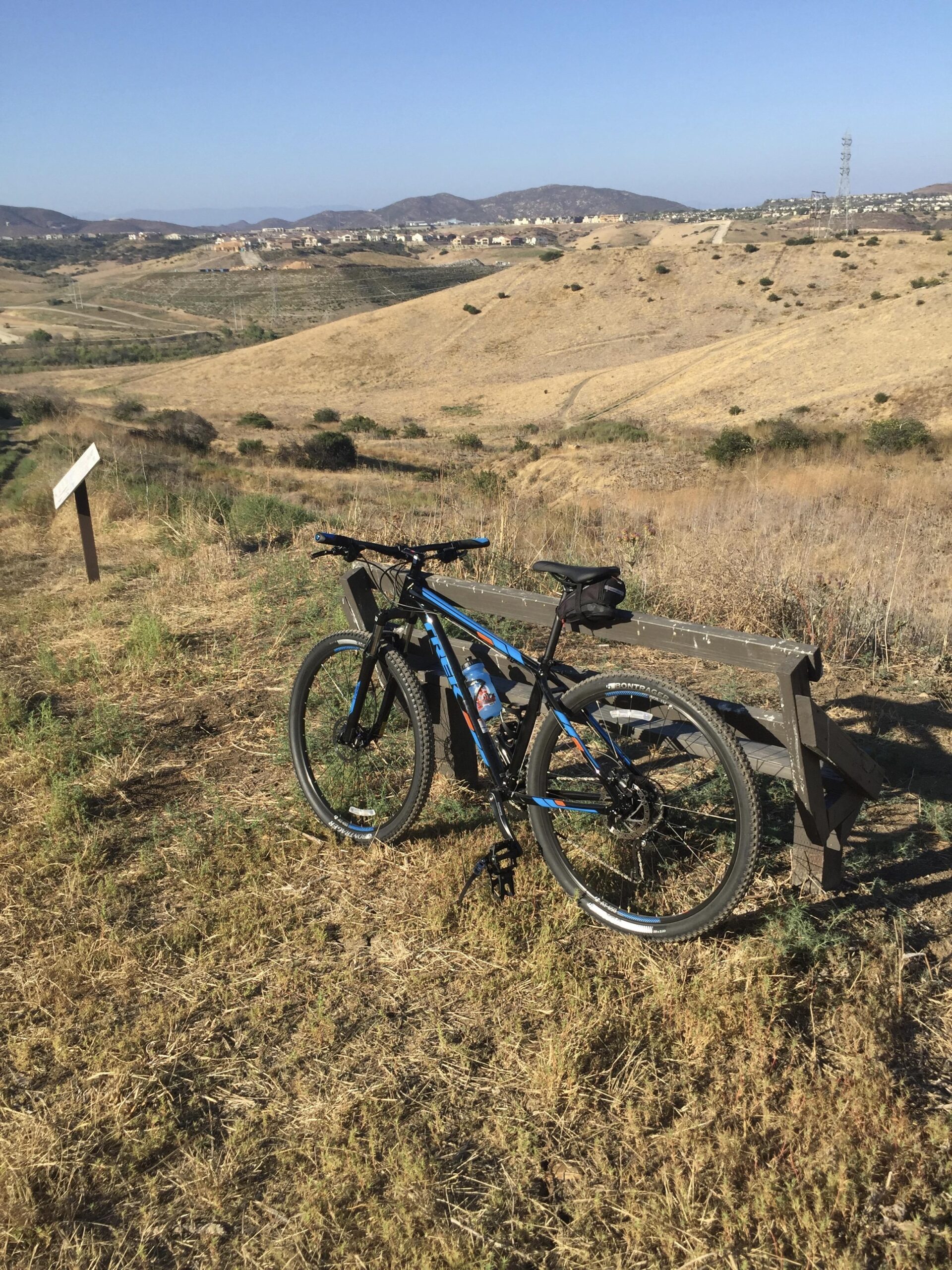 Trek Marlin 7: A mountain bike rests against a wooden bench on a hillside, with dry grass and sparse vegetation surrounding it. In the background, rolling hills extend into the distance, dotted with houses and under a clear blue sky. A pathway meanders through the landscape, suggesting potential biking trails.